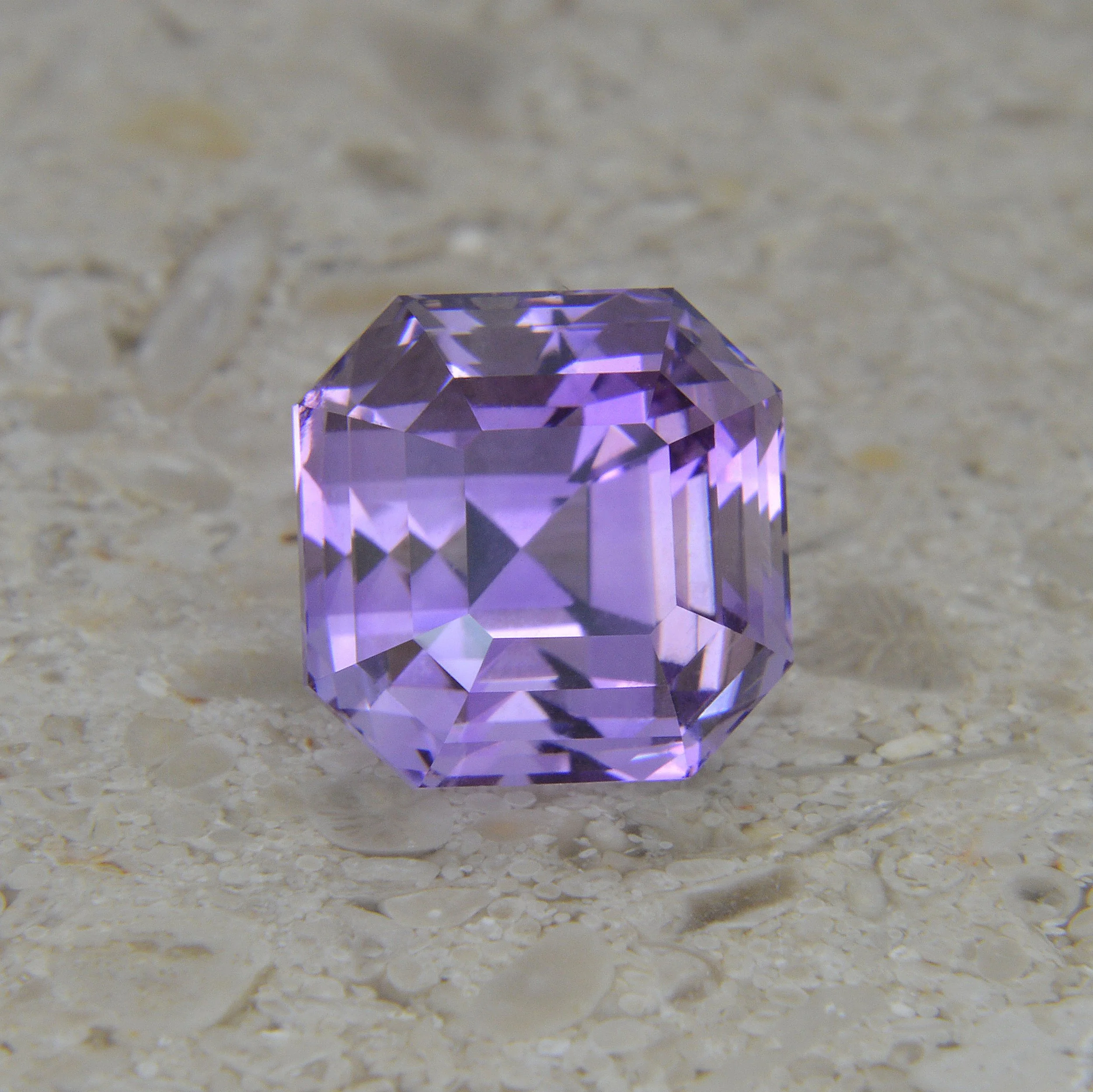 A purple, faceted gemstone with an octagonal shape resting on a textured beige surface.