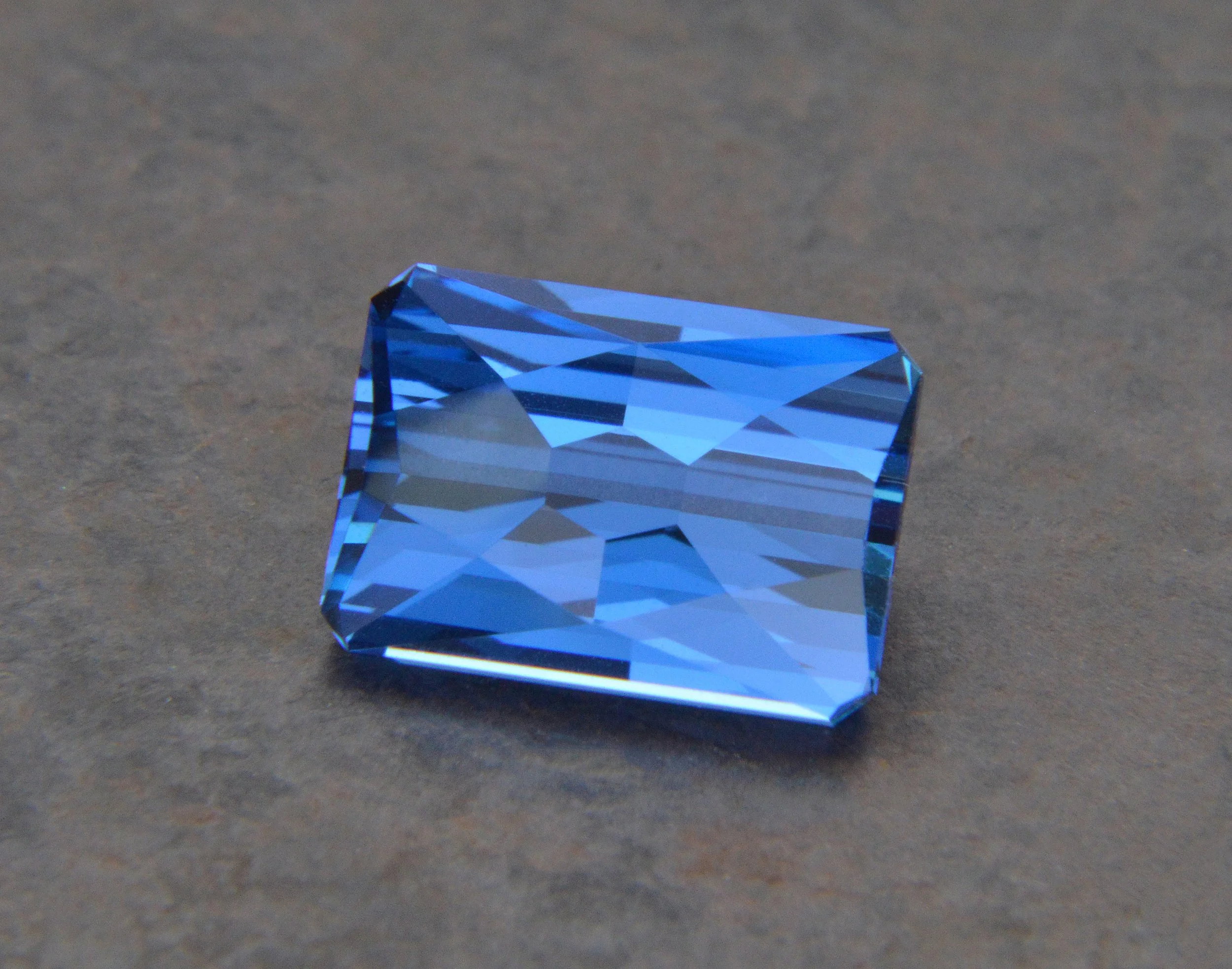 A blue square-cut gemstone, possibly a sapphire, resting on a textured surface.