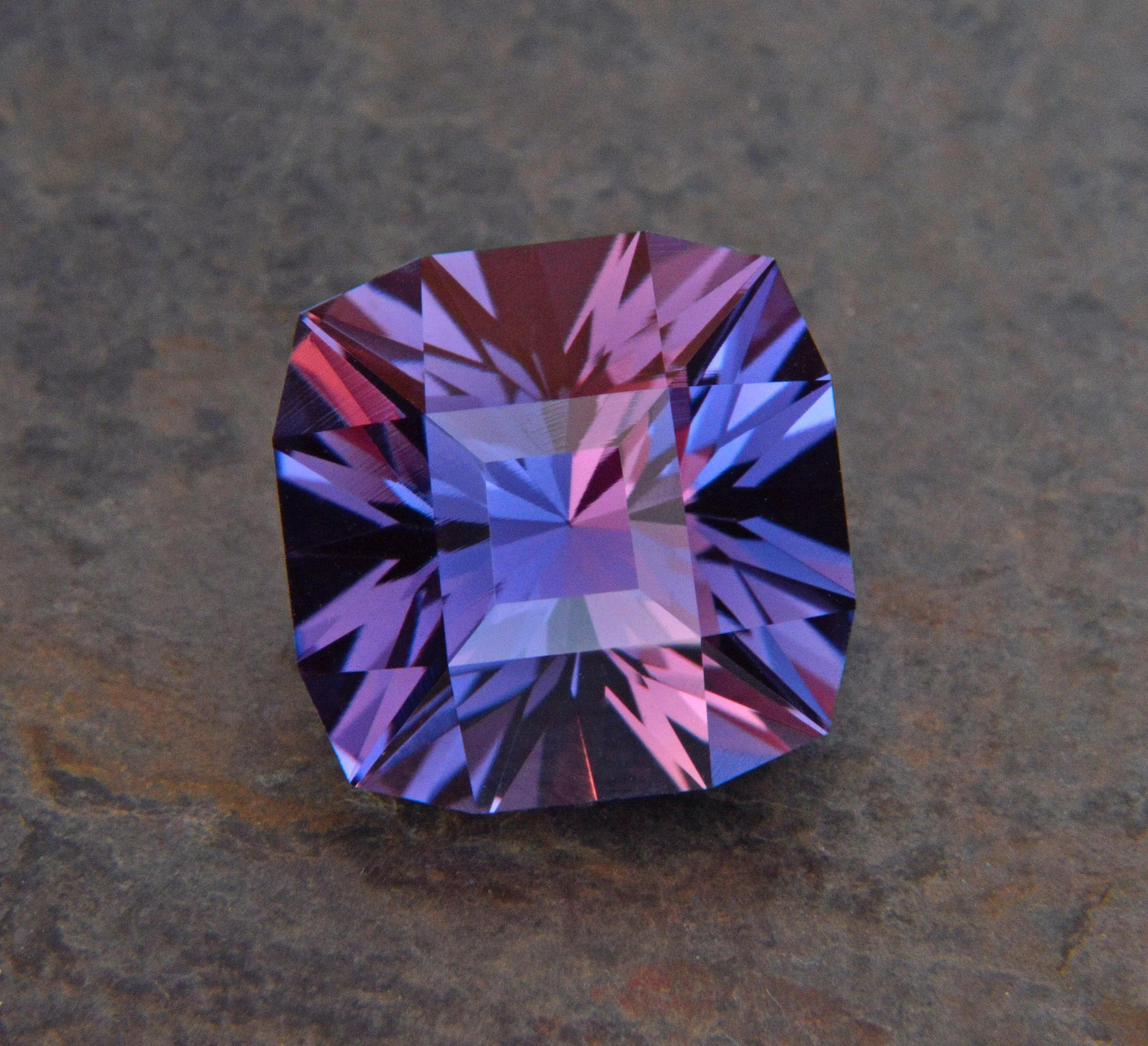 A large, square-cut, purple gemstone with intricate facets, resting on a textured dark surface.
