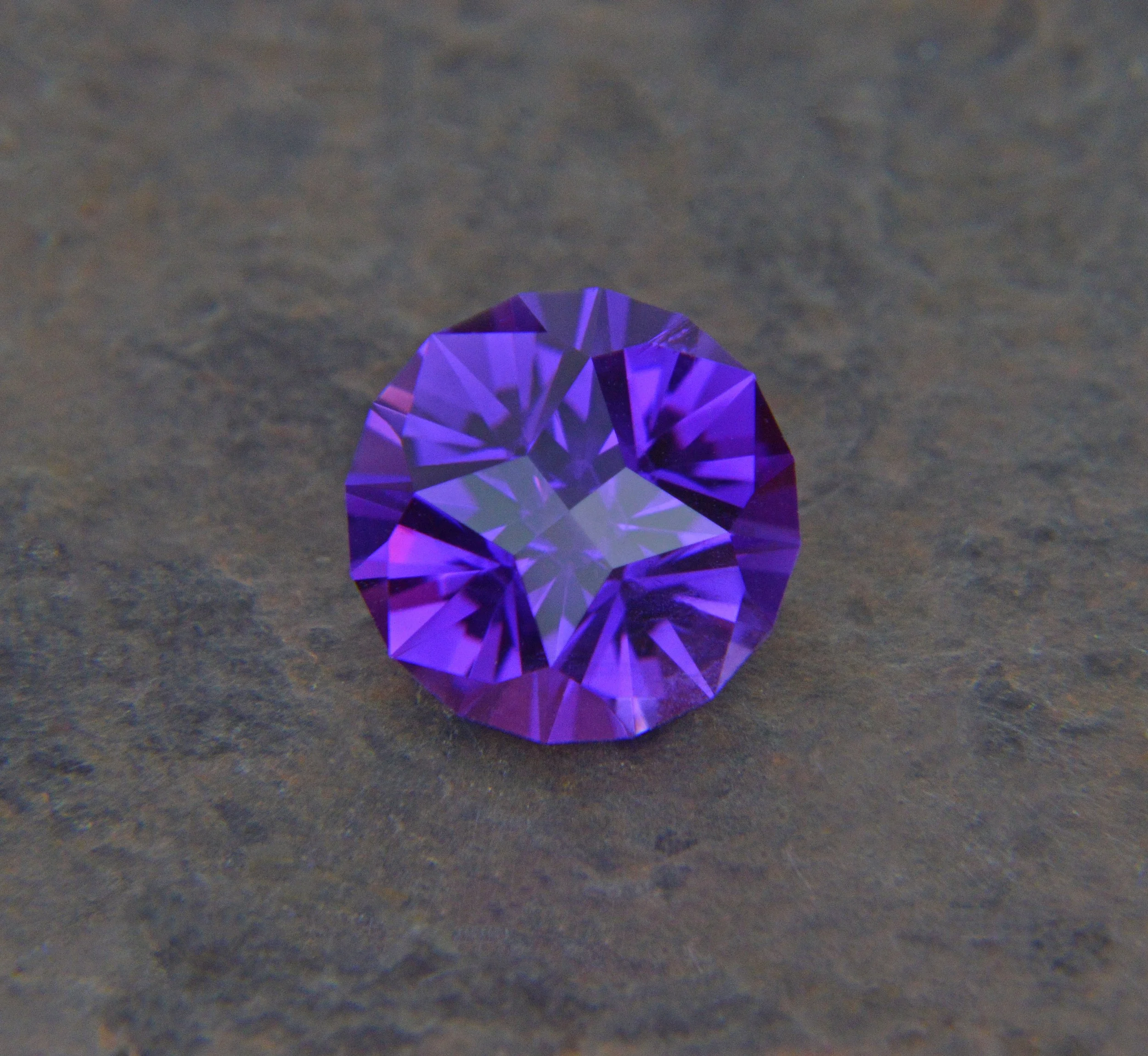 Close-up of a purple gemstone with a round cut on a gray surface.