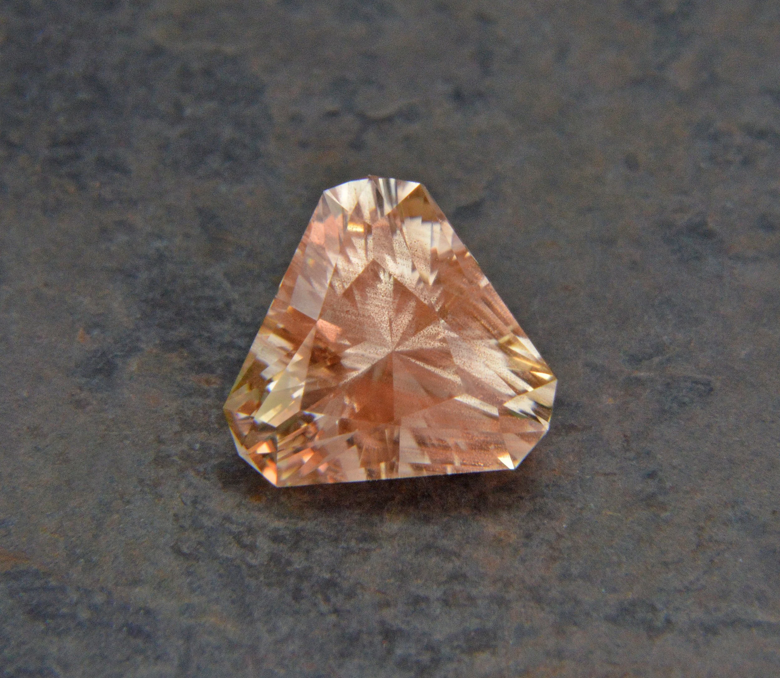 A faceted gemstone with a pale pink or peach color, placed on a dark gray surface.