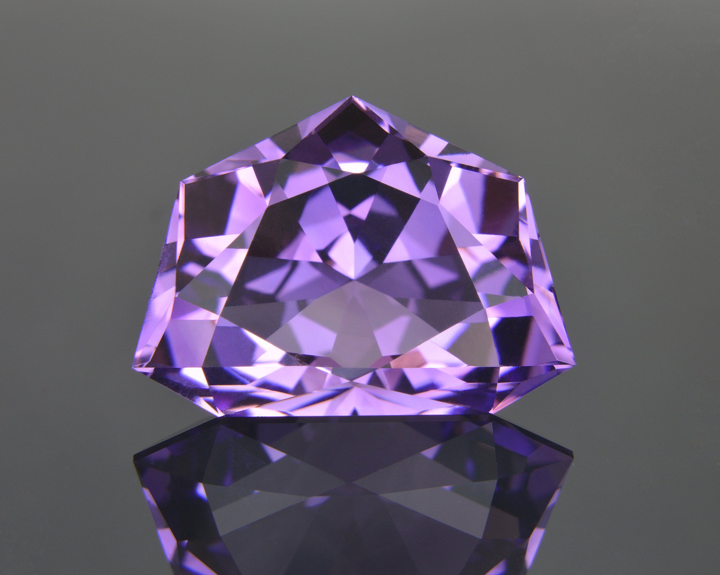 A purple gemstone with multiple facets, resting on a reflective surface, with a neutral gray background.