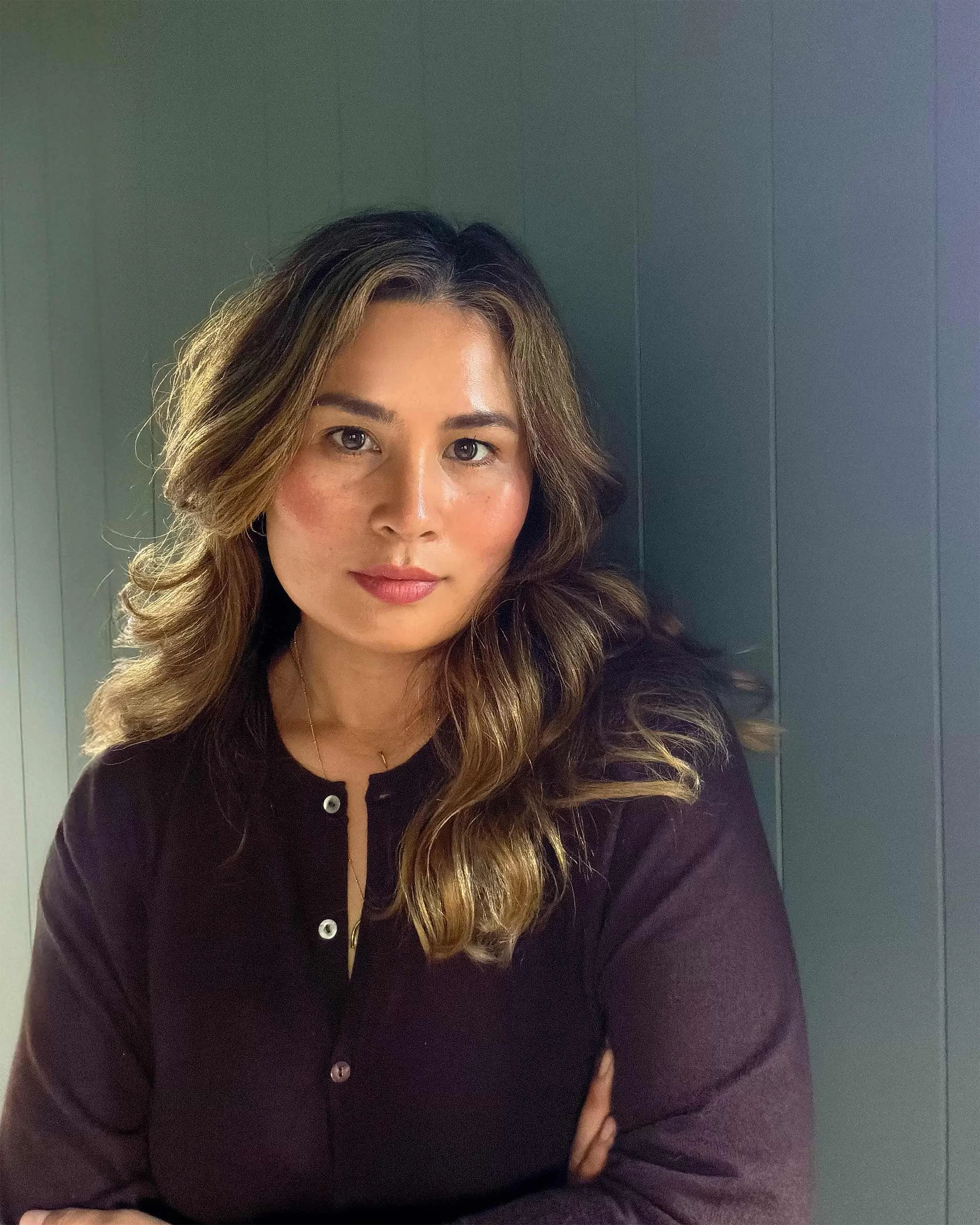 A woman with wavy brown hair and light skin, wearing a black top, standing against a dark gray paneled wall, looking directly at the camera.
