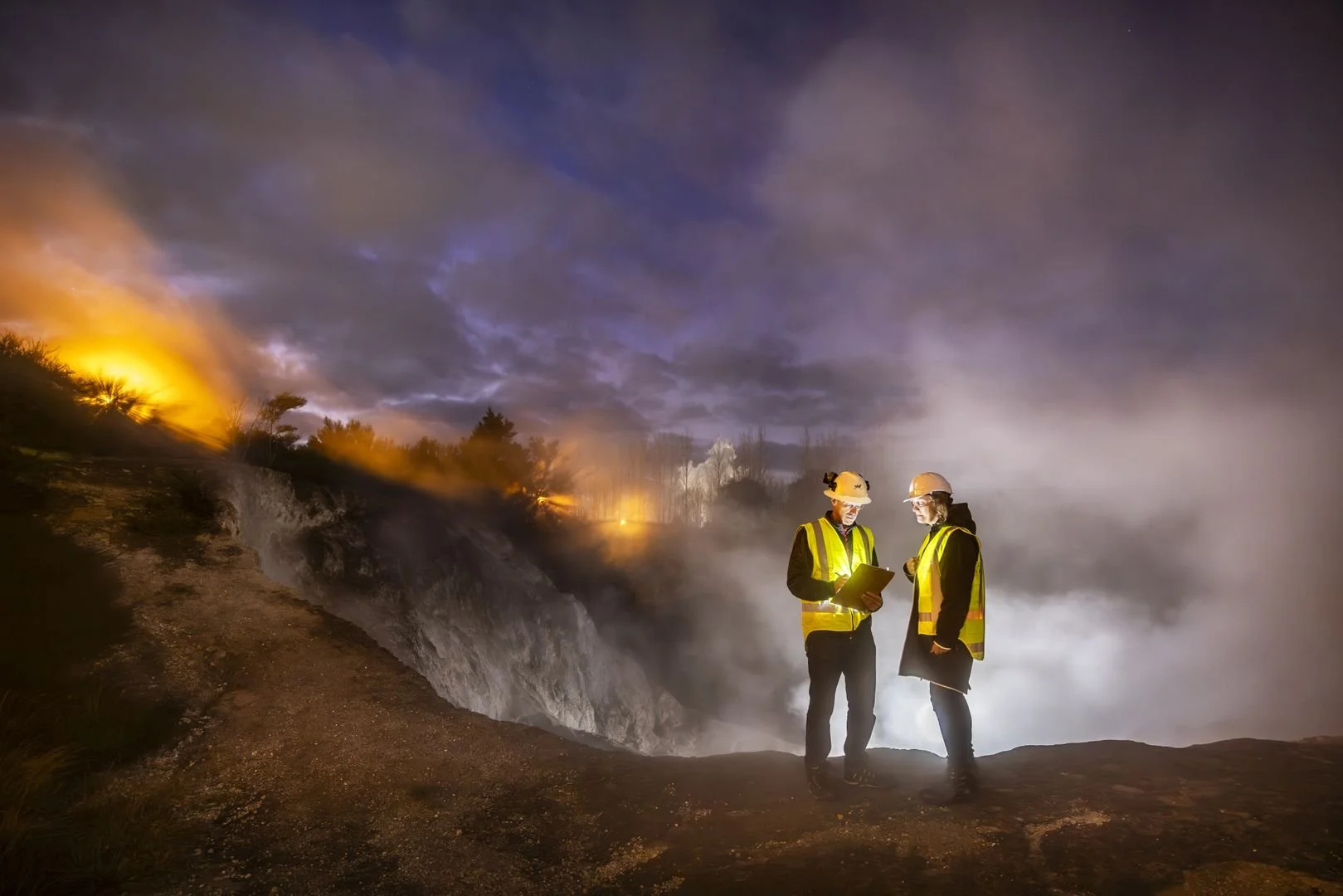 Two peopl, man and woman, stand on the edge of a geothermal pit, smoke billowing in the air behind them.
