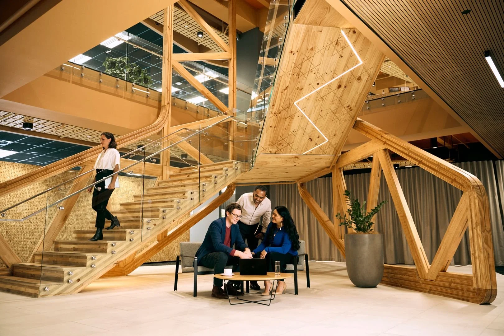 People in a modern office lobby with a large, intricate wooden staircase and a wood and glass railing. Two people are seated on a small couch, talking and looking at a laptop on a coffee table, while a third person stands nearby. Another person is descending the staircase.