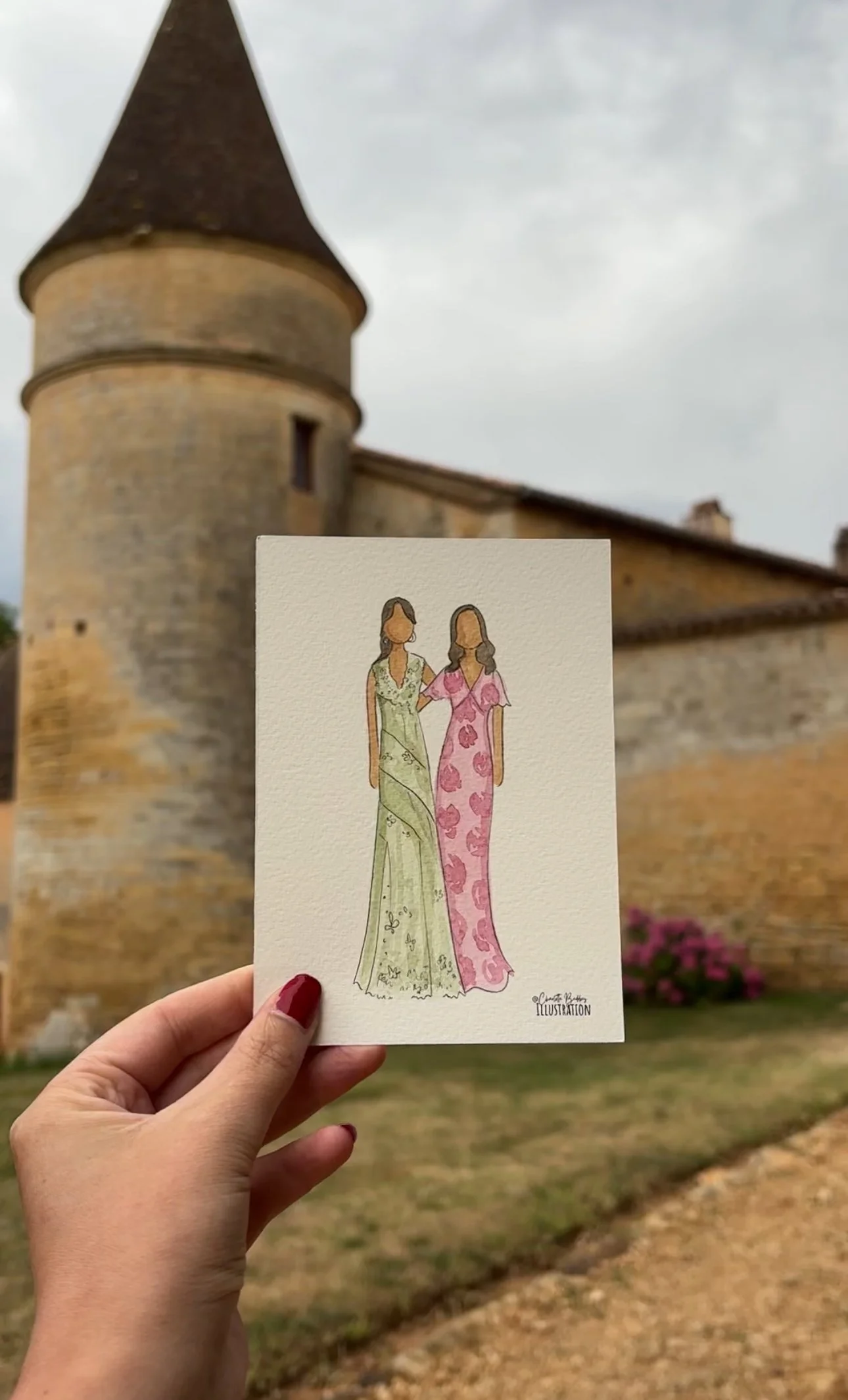 A hand holding a watercolor illustration of two women in elegant dresses in front of a historic stone castle with a turret.