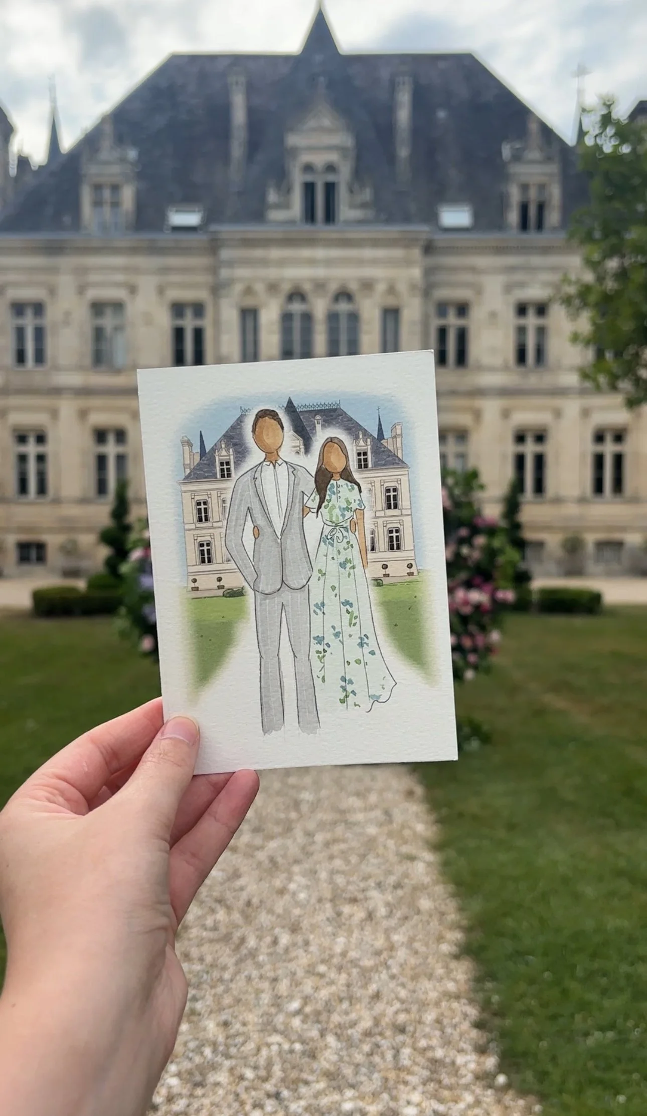 Hand holding a watercolor illustration of a couple in wedding attire in front of a historic mansion, with well-manicured gardens and a gravel pathway.