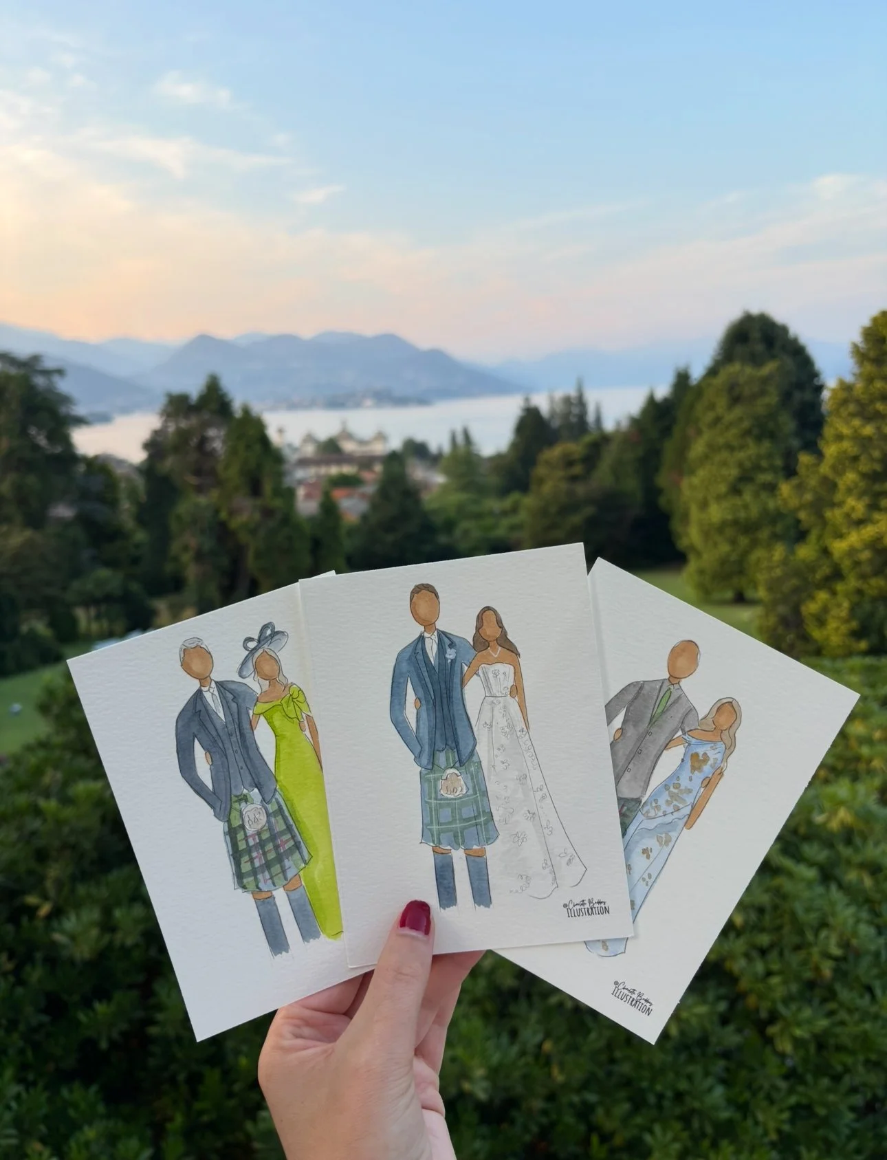A hand holding three illustrated paper cards featuring fashion designs of men and women in stylish outfits, with a scenic outdoor background of trees, mountains, and a lake during sunset.