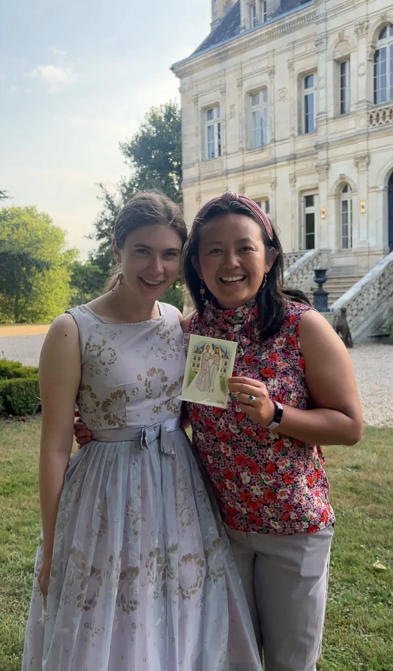 Two women standing outdoors in front of a historic building, smiling, one holding a religious card. One woman is wearing a light-colored floral dress, and the other is wearing a sleeveless floral top and light-colored pants.