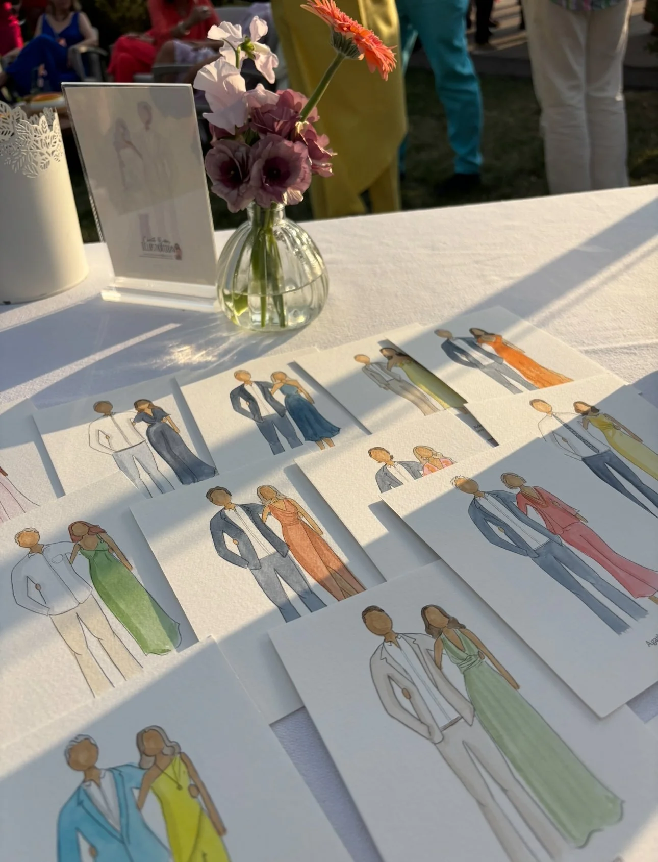 Sketches of couples in formal attire, displayed on a table with sunlight, a vase with pink and white flowers, and a sign, at an outdoor event.
