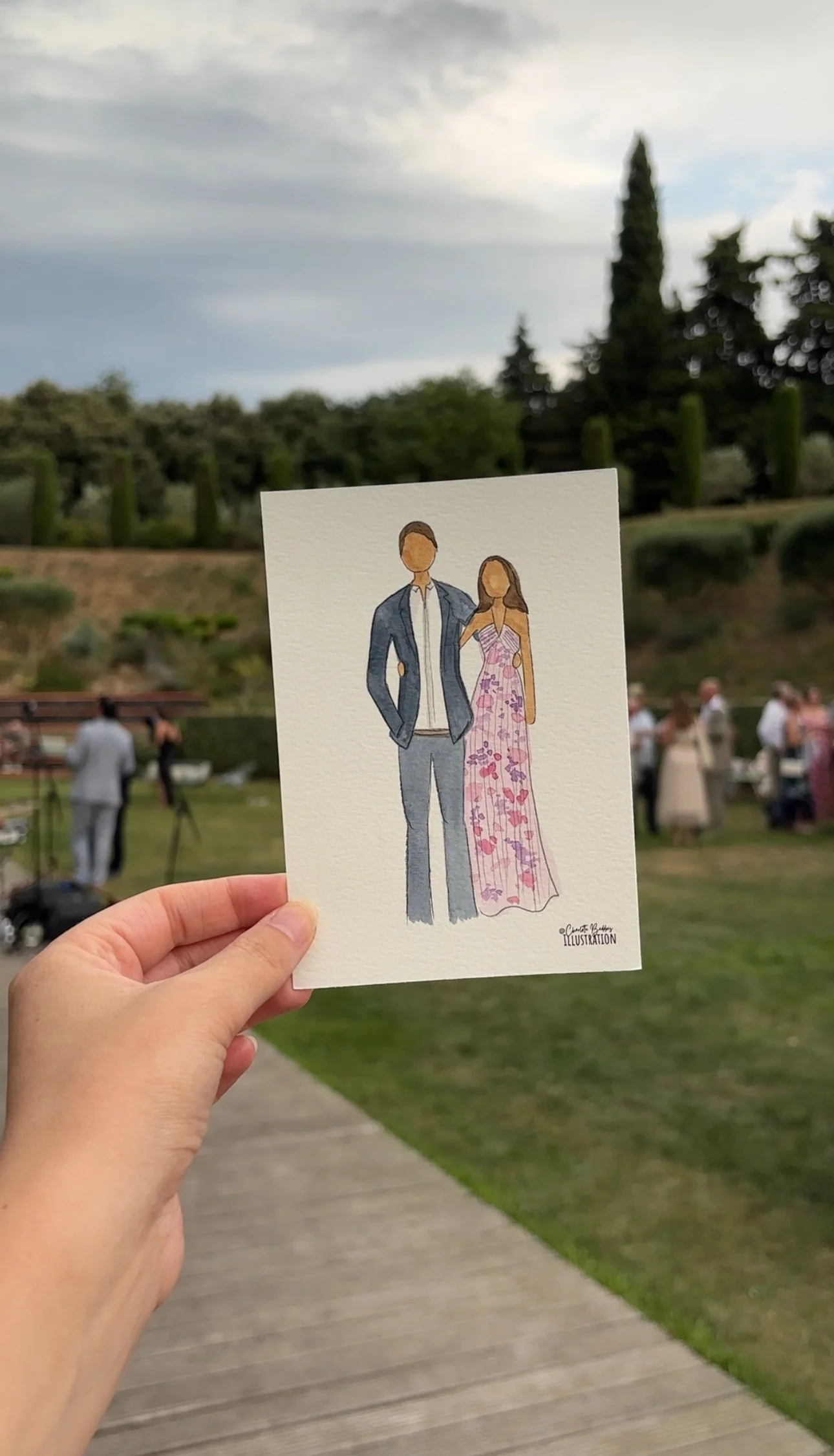 A hand holding a watercolor illustration of a couple dressed for a formal event, with a garden or park setting with people in the background.