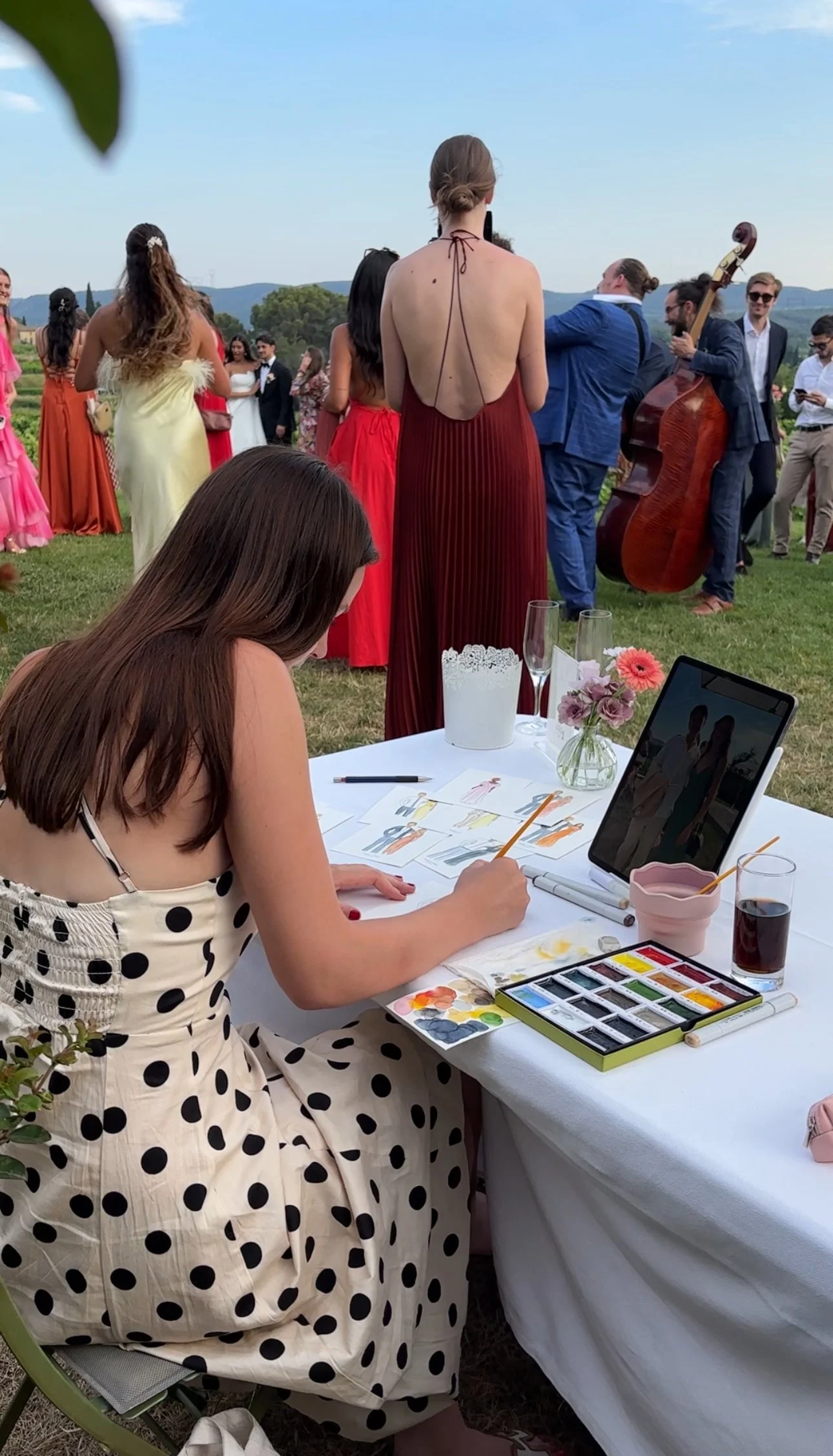 A woman in a polka dot dress sitting at a table with watercolor paints and an iPad, painting at an outdoor event with people dancing and playing music in the background.