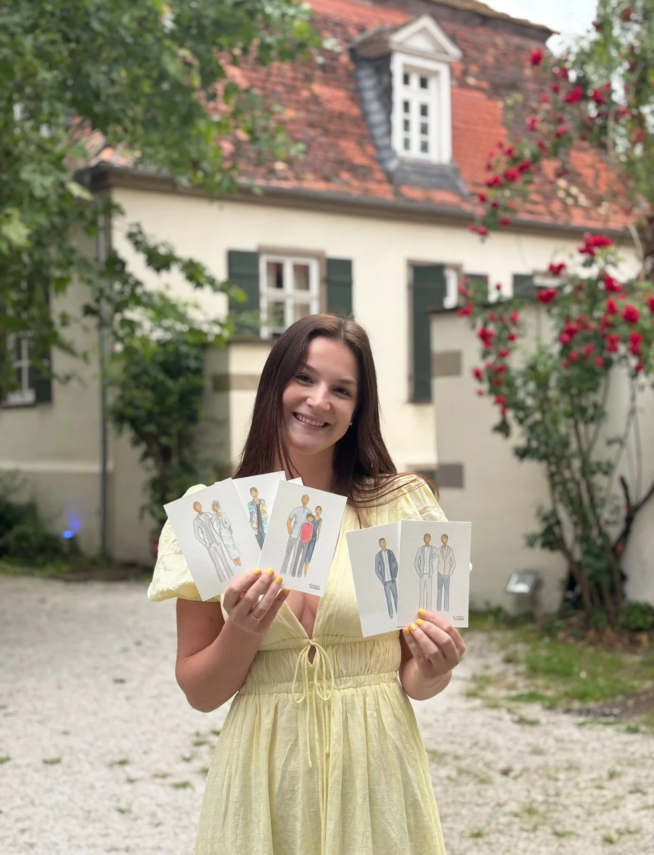 A woman in a yellow dress smiling and holding illustrated cards of various fashion styles in an outdoor garden with trees, pink flowers, and a white house with green shutters in the background.