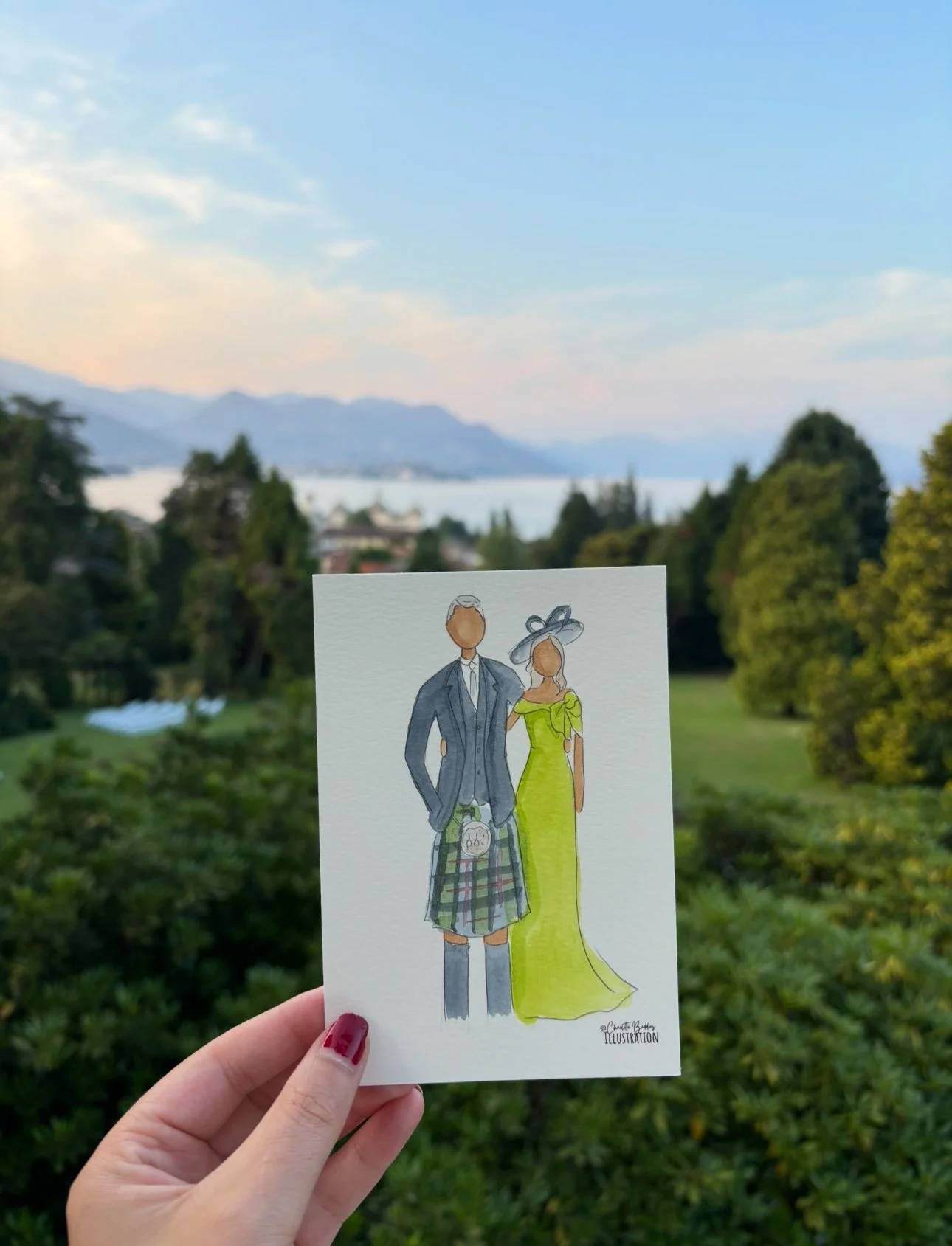 A hand holding a drawing of a man and woman in formal attire, with scenic greenery, trees, a body of water, and mountains in the background.