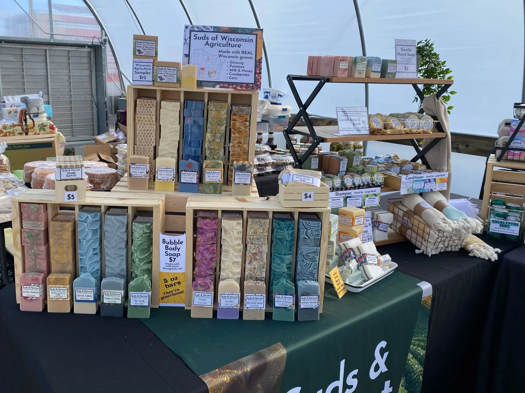 Display of handmade soap bars and skincare products at a market stall, labeled 'Suds of Wisconsin Agriculture', featuring different colors, shapes, and sizes of soap, along with signs indicating prices and product details.