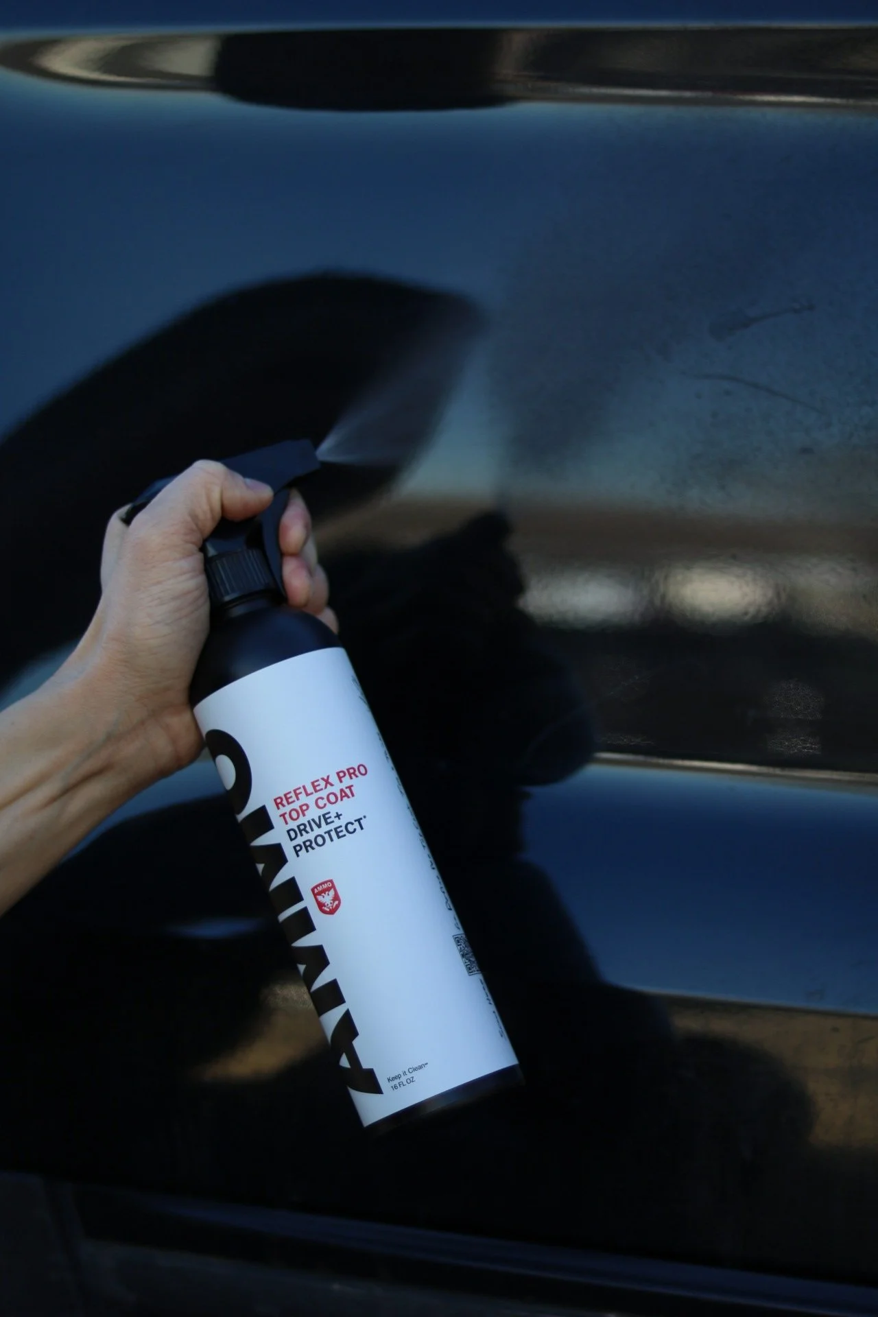 A person applying a spray bottle labeled 'Reflex Pro Top Coat' on the surface of a black car.
