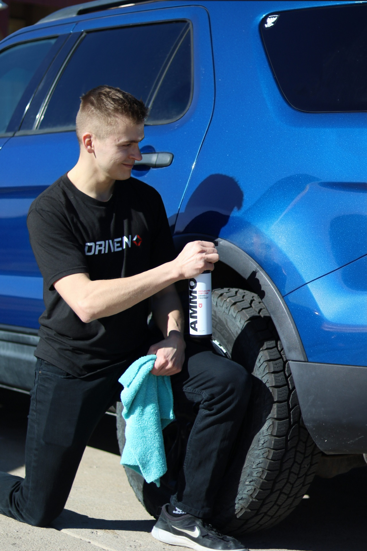 A young man kneeling beside a blue vehicle, applying tire coating with a spray can, holding a blue cloth in his other hand.
