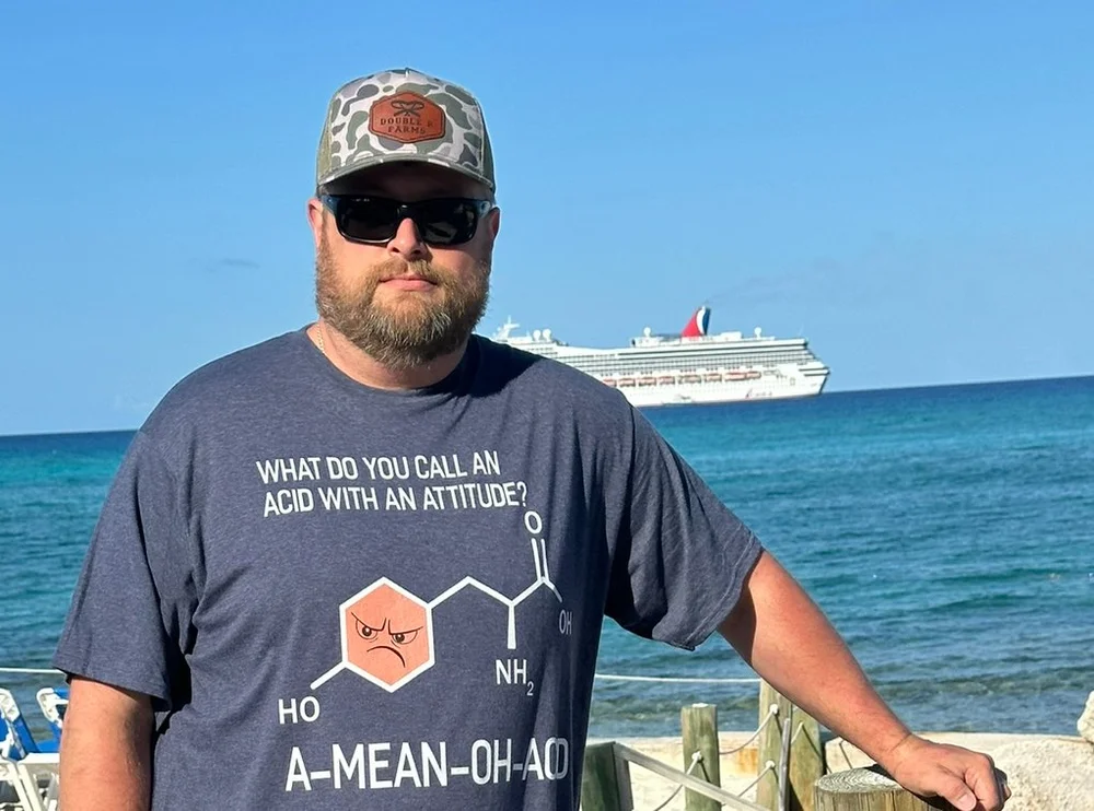 Man wearing sunglasses and a cap, standing by the sea with a cruise ship in the background.