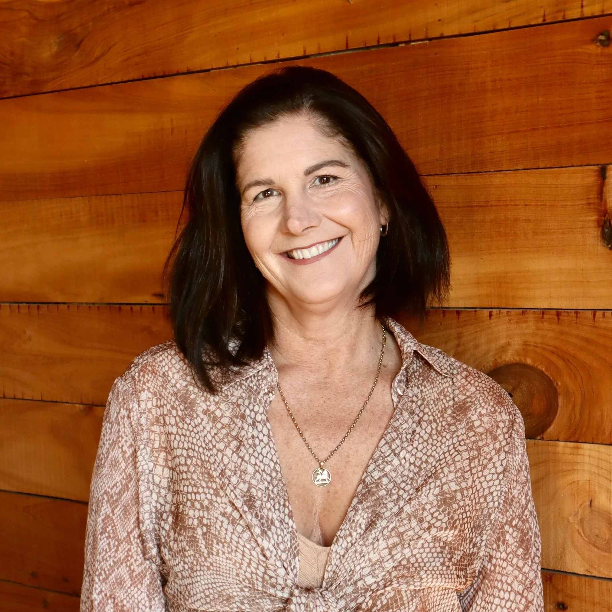 Smiling woman with shoulder-length dark hair, wearing a patterned blouse, a necklace, and earrings, standing against a wooden wall background. Maree Coombes.
