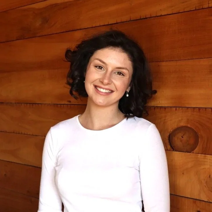 A young woman with short, curly dark hair smiling, wearing a white top and pearl earrings, standing in front of a wooden wall.
Sienna Arnold