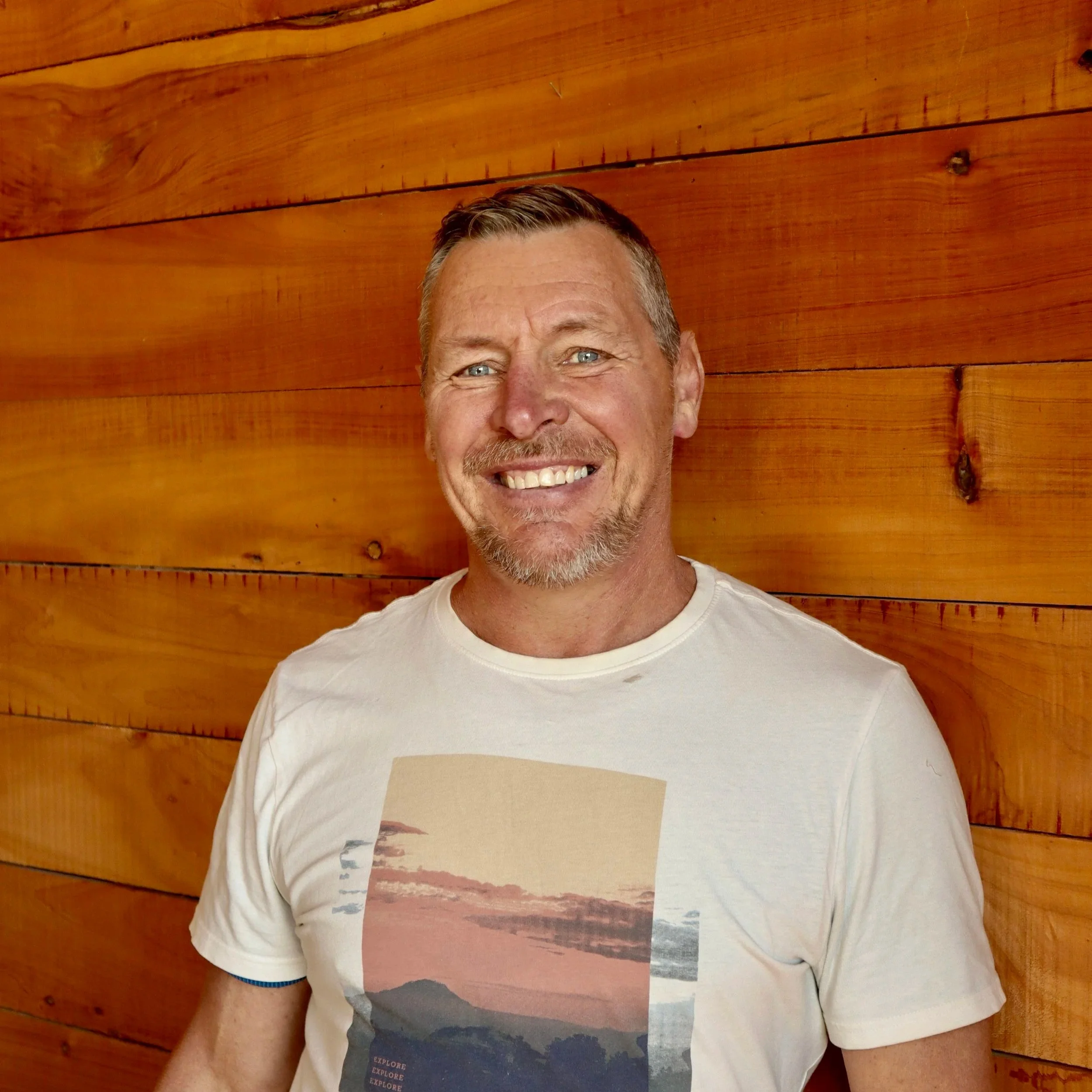 A middle-aged man with short, light brown hair and a beard, smiling, standing in front of a wooden wall. He is wearing a white T-shirt with a graphic of a sunset landscape. Phillip Coombes.