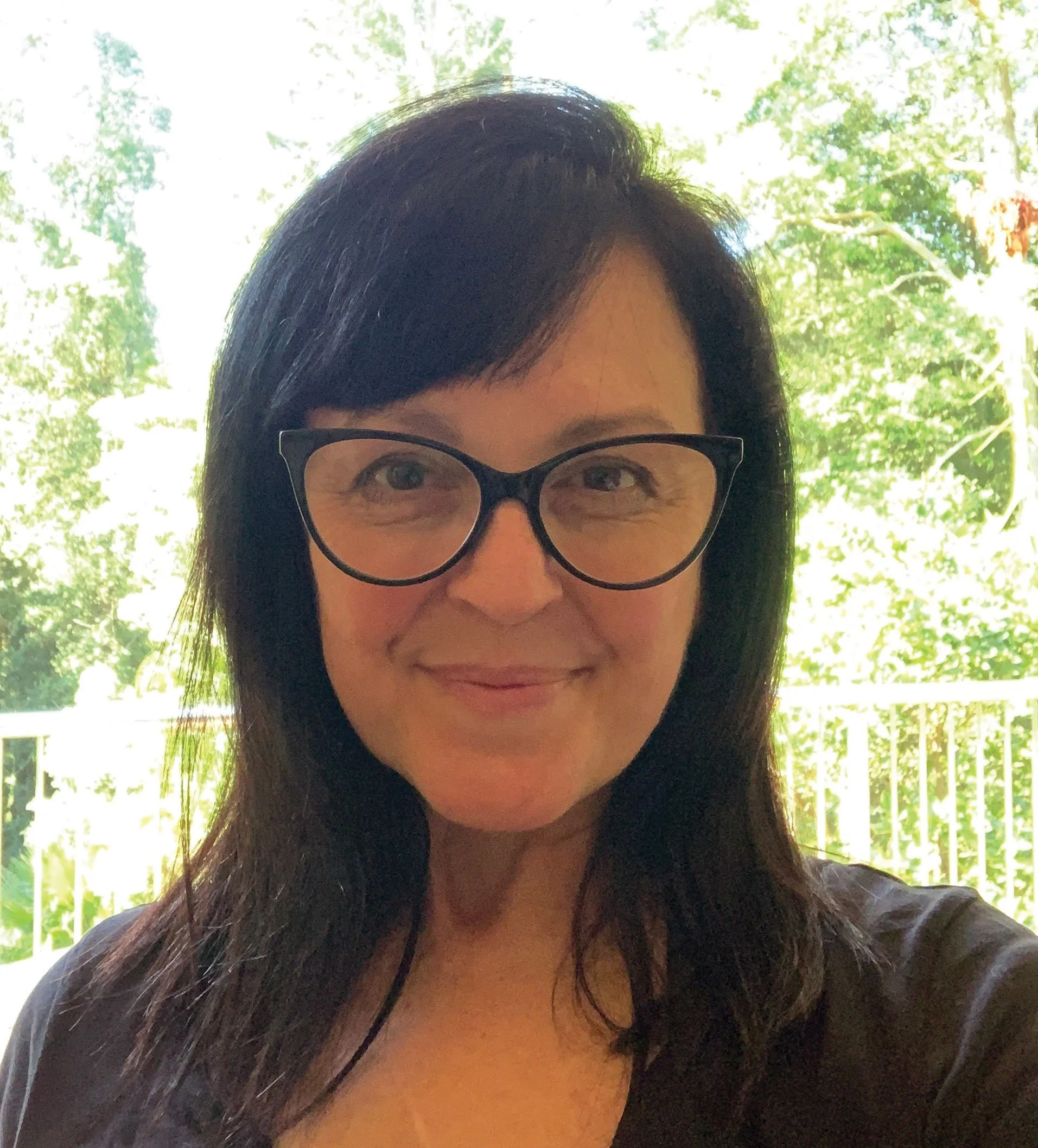 A woman with black hair, wearing glasses, smiling outdoors with trees and greenery in the background.