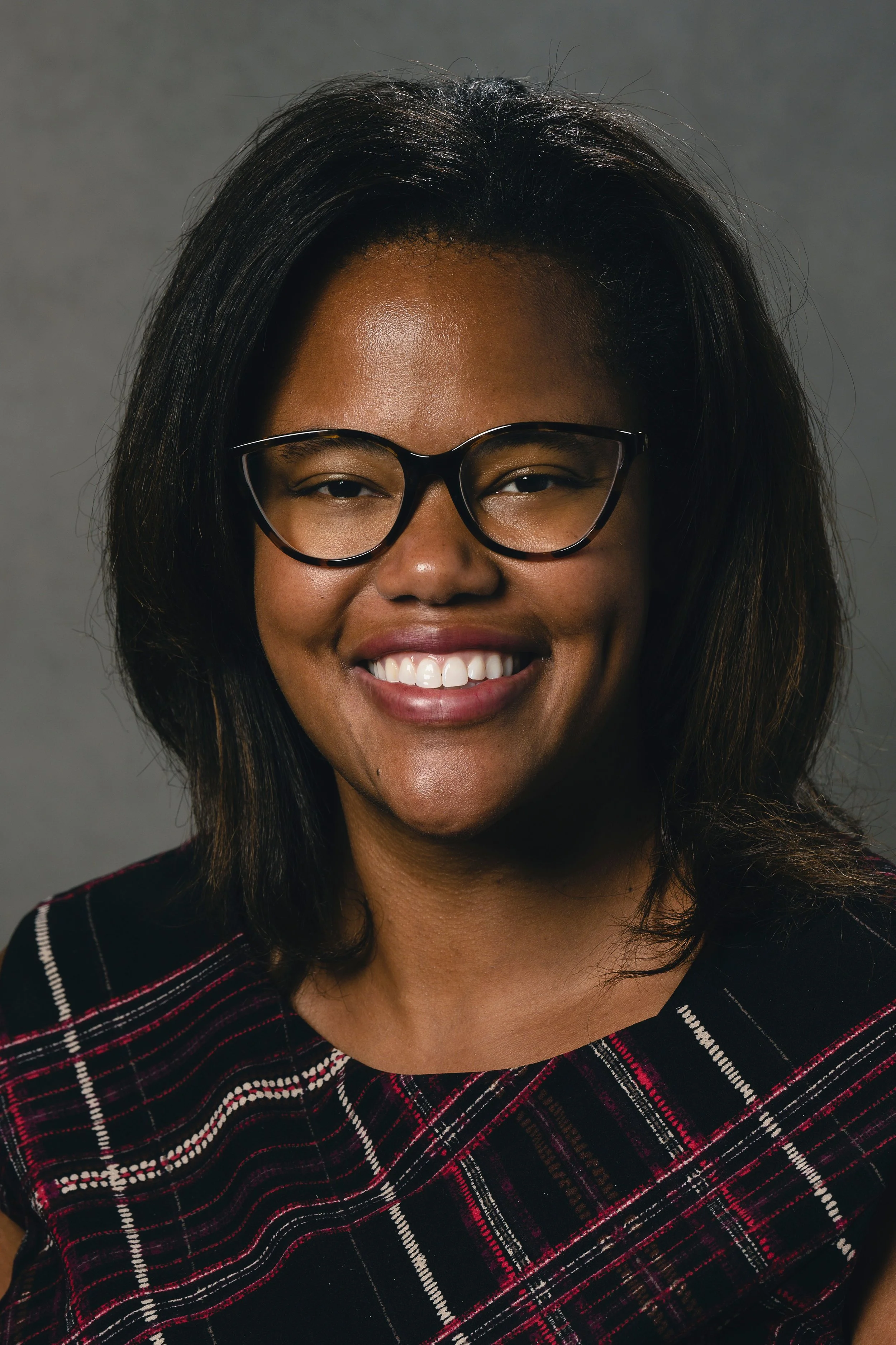 A woman with dark hair, wearing glasses, smiling against a gray background.