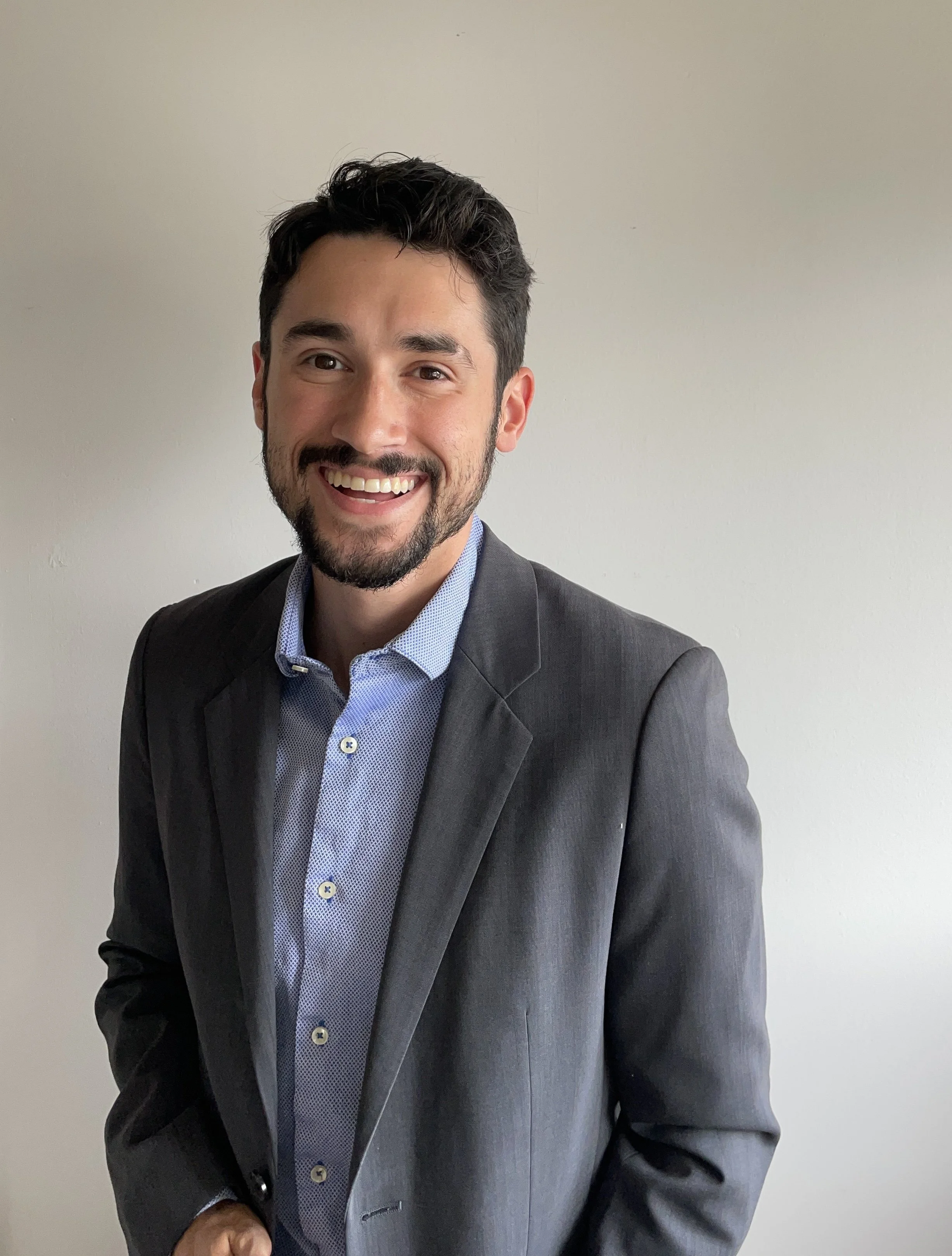 A smiling man in a gray suit and blue dress shirt standing against a plain light-colored wall.