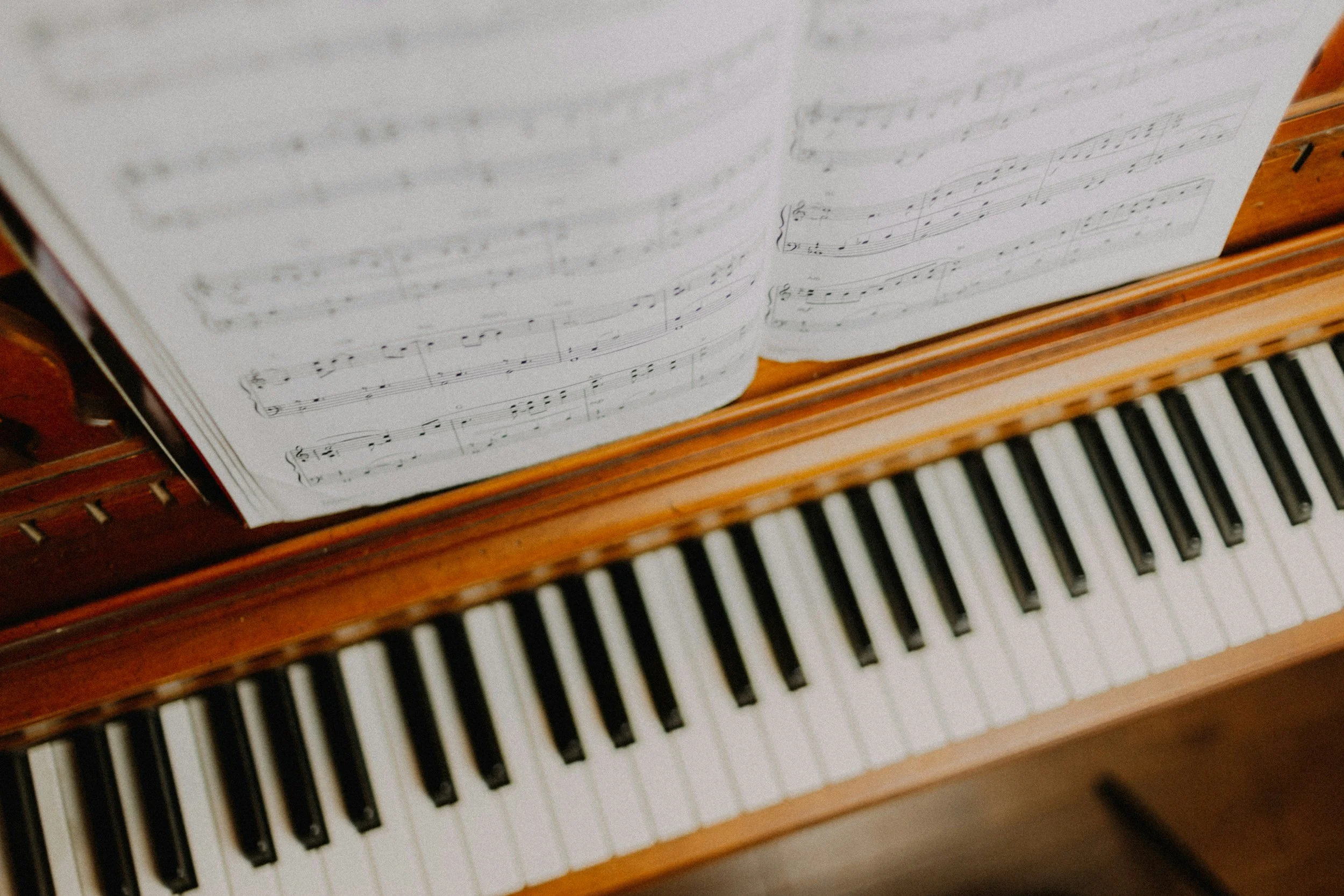 Close-up of a sheet music book resting on a wooden keyboard of a piano, showing printed musical notation and black-and-white keys.