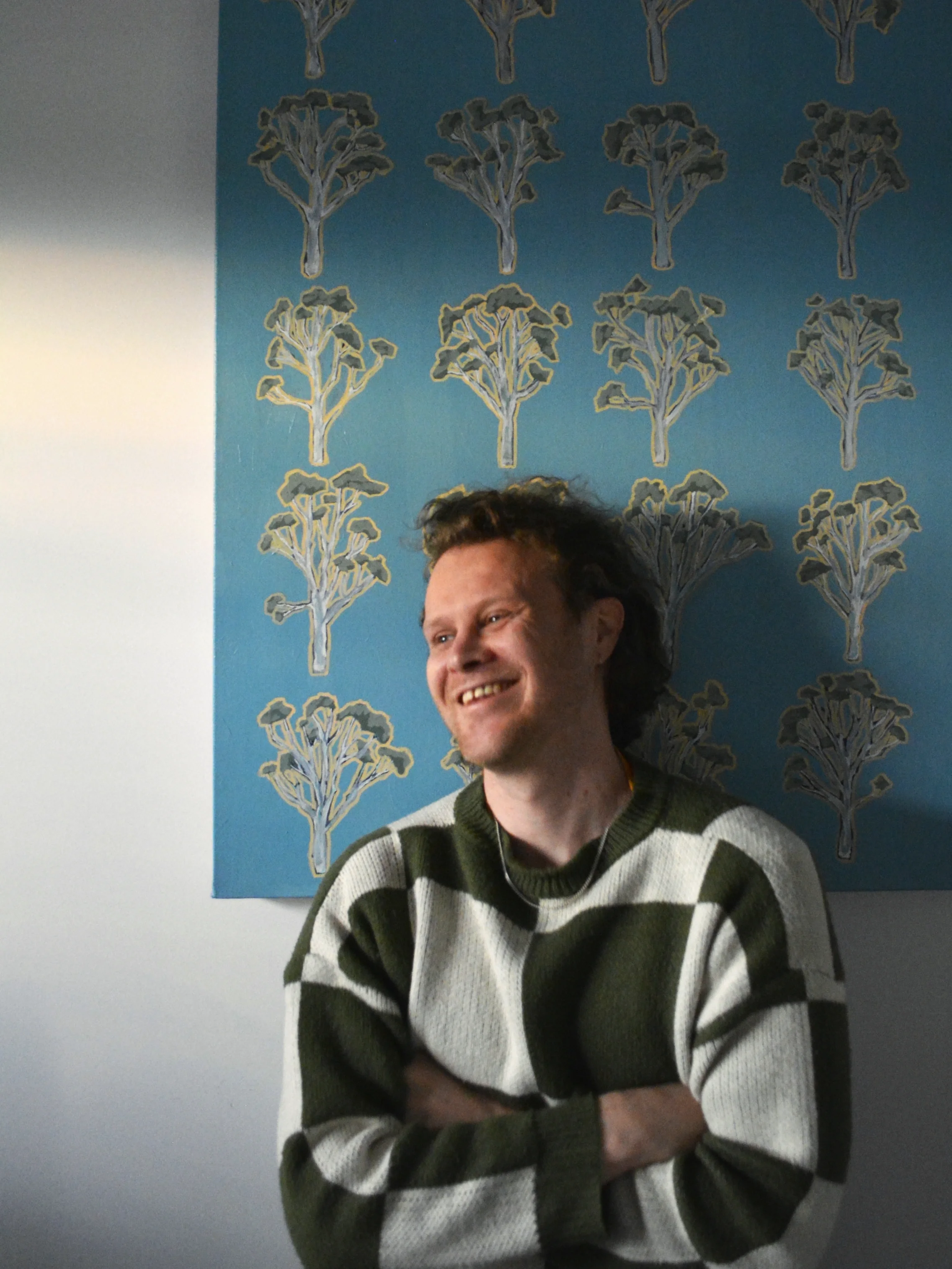 A man with brown curly hair wearing a green and white striped sweater, smiling with arms crossed, standing in front of a wall with blue wallpaper decorated with tree patterns.