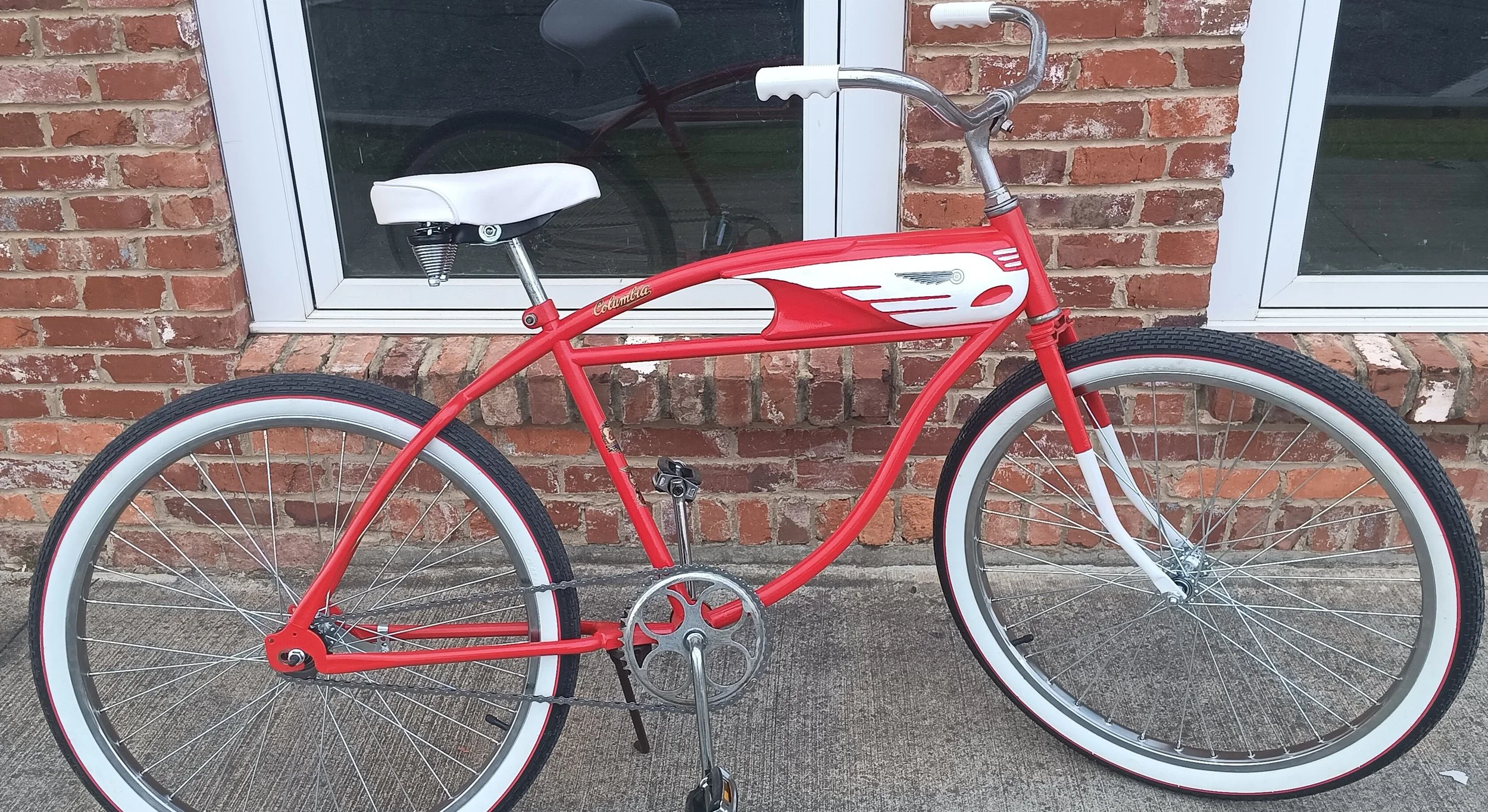 Red and white vintage-style bicycle with a white seat and white-walled tires, parked in front of a brick wall and a window.