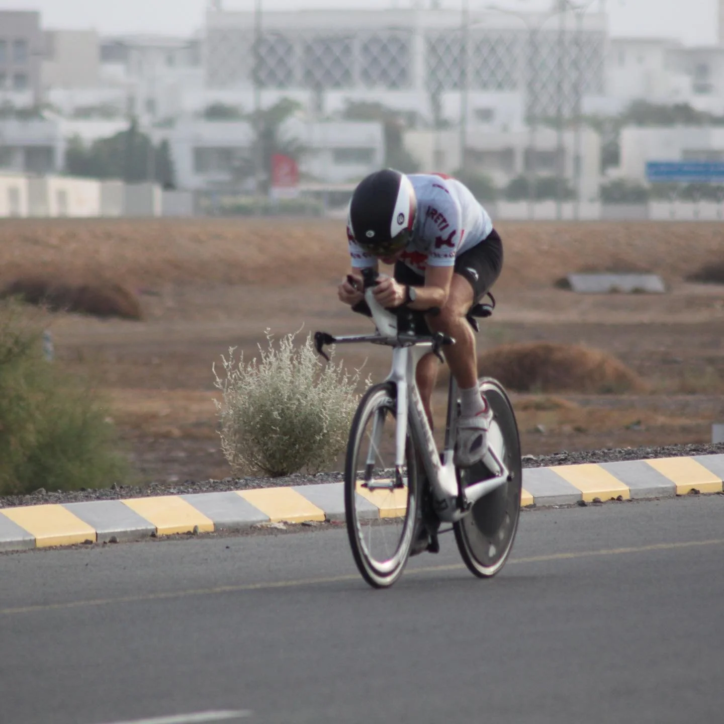 A cyclist wearing a helmet and sports attire rides a time trial bike on a paved road in an arid area with bushes and buildings in the background.