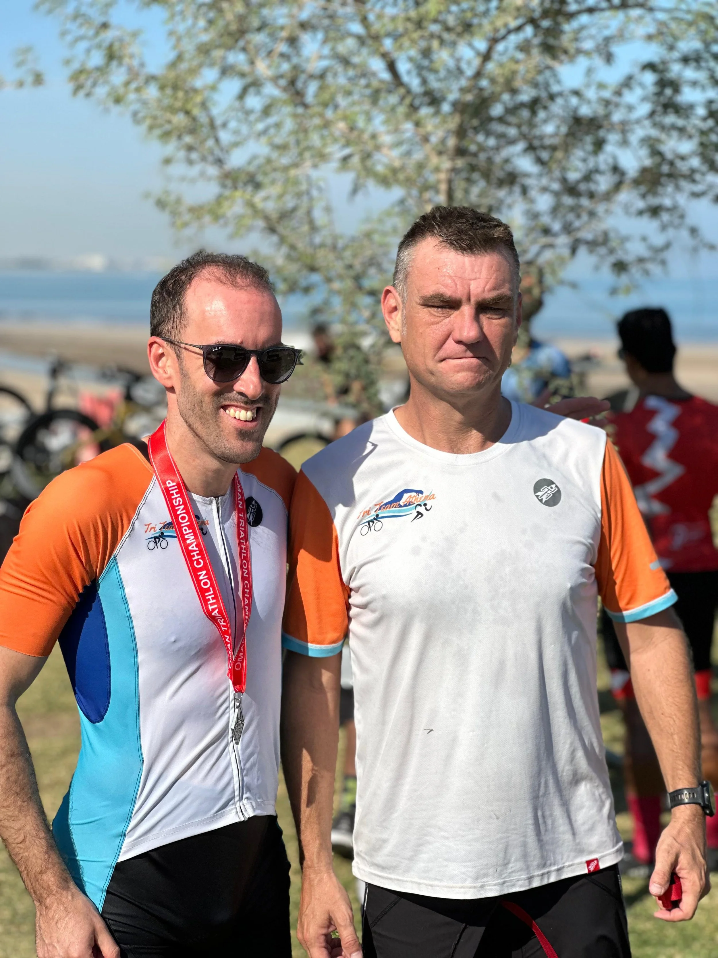 Two men in athletic gear standing outdoors near trees with a beach and water in the background. One man is smiling and wearing sunglasses, a triathlon medal around his neck, and a colorful athletic shirt. The other man is looking serious, wearing a white and orange athletic shirt, and has a fitness tracker on his wrist.