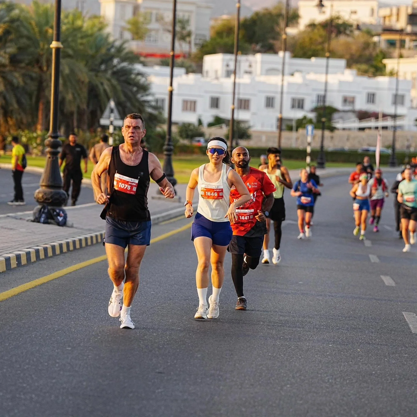 People running in a marathon on a city street during sunset, with runners wearing numbered bibs and athletic clothing.