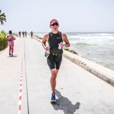 Female runner in black athletic wear and pink cap running along a beachside path with ocean waves in the background, other runners in the distance.