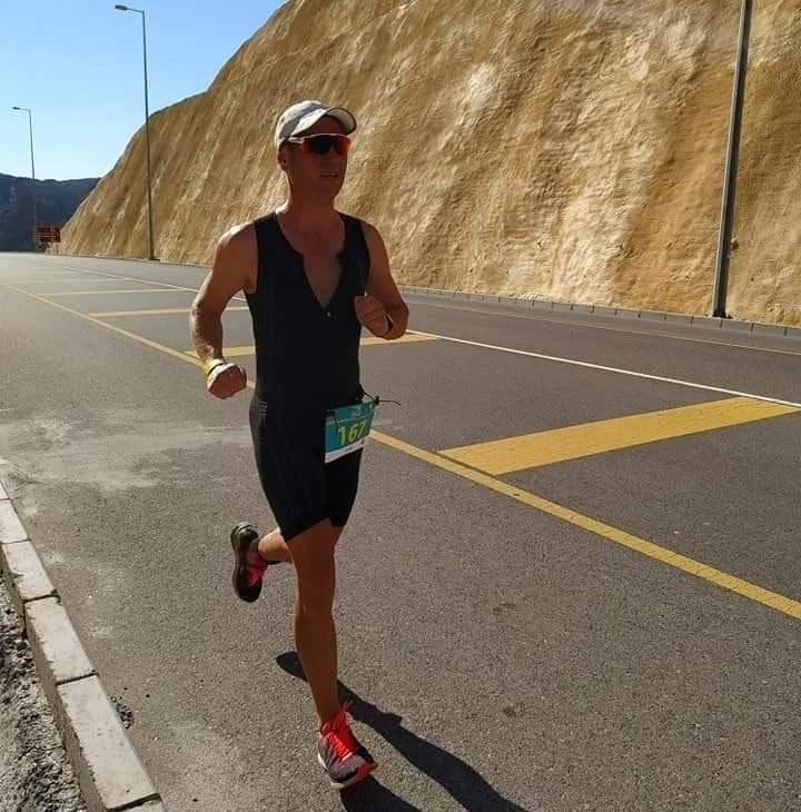 A man running on a paved road next to a rocky yellow cliff, wearing sunglasses, a hat, a sleeveless black top, black shorts, and red running shoes.