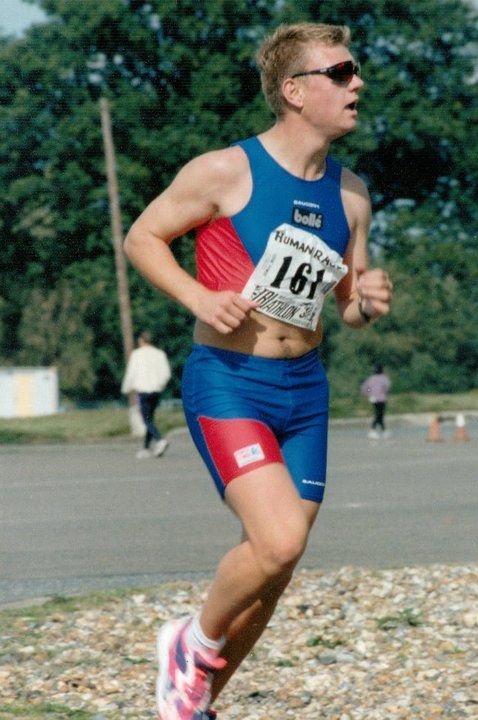 A male runner in a blue and red athletic outfit running outdoors during a race, wearing sunglasses and a race bib number 164.