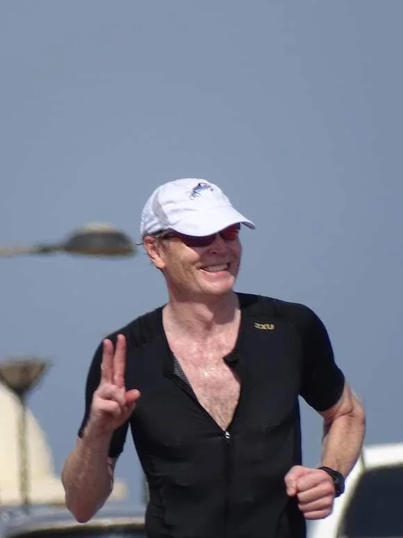 A man wearing a white cap, sunglasses, and a black athletic shirt running outdoors, smiling and making a peace sign with his right hand.