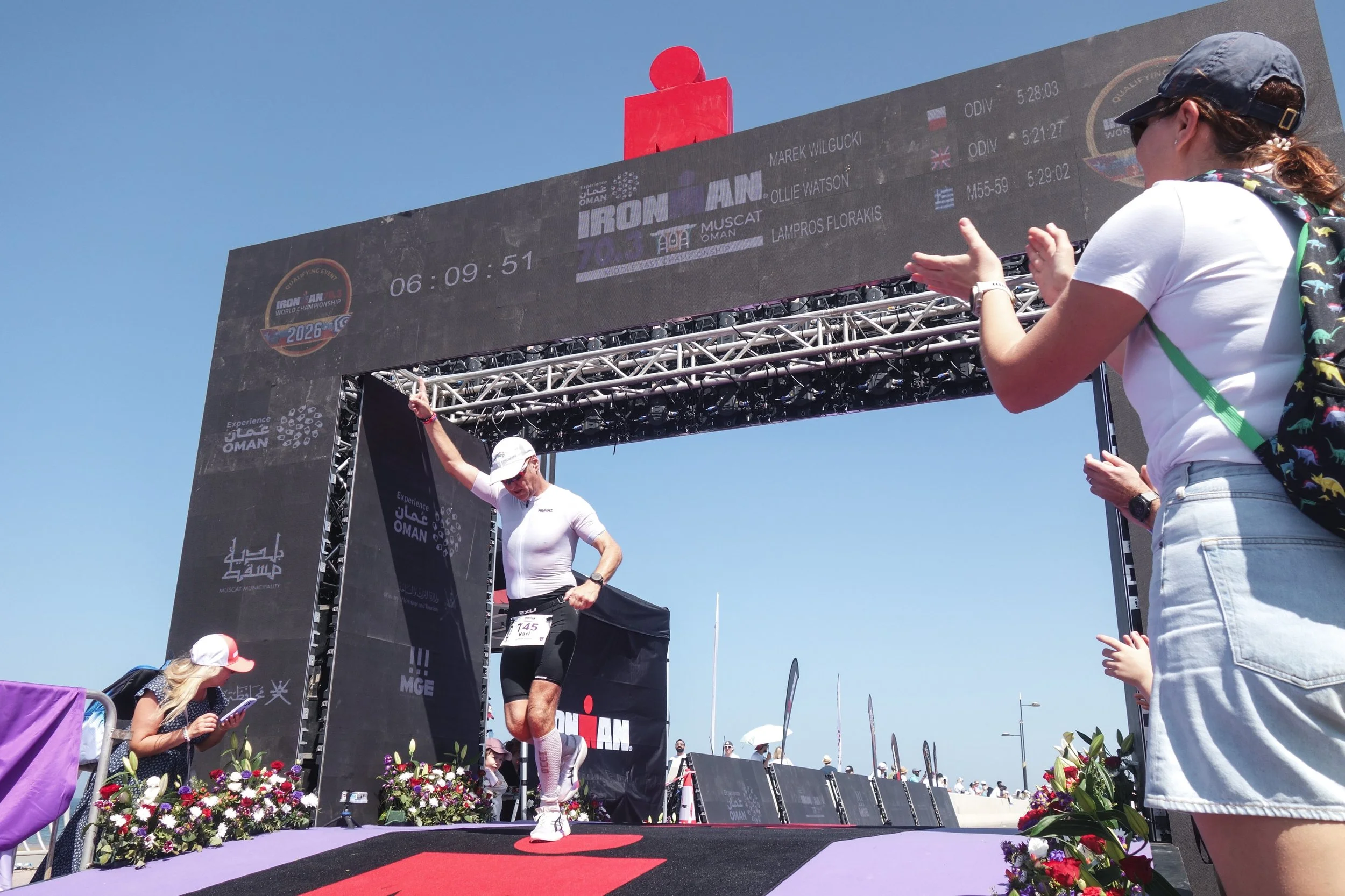 A male athlete crossing the finish line at an ironman triathlon event, with spectators clapping and cheering, under a large digital timer and event signage during daytime.
