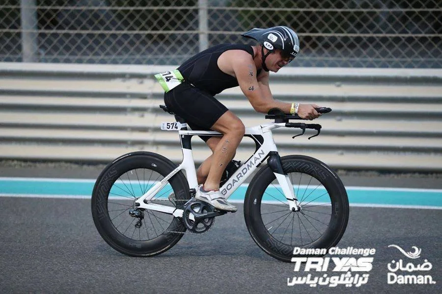 A cyclist riding a white Boardman triathlon bike at a race track, wearing a black helmet and black athletic attire, with race bib number 574, during the Daman Challenge Triyas event.