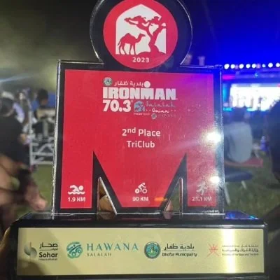 A clear plastic award plaque for the 2023 IRONMAN 70.3 race, indicating 2nd place finish by TriClub. The award is displayed outdoors with people and a lit stage in the background, and it features logos and awards information.