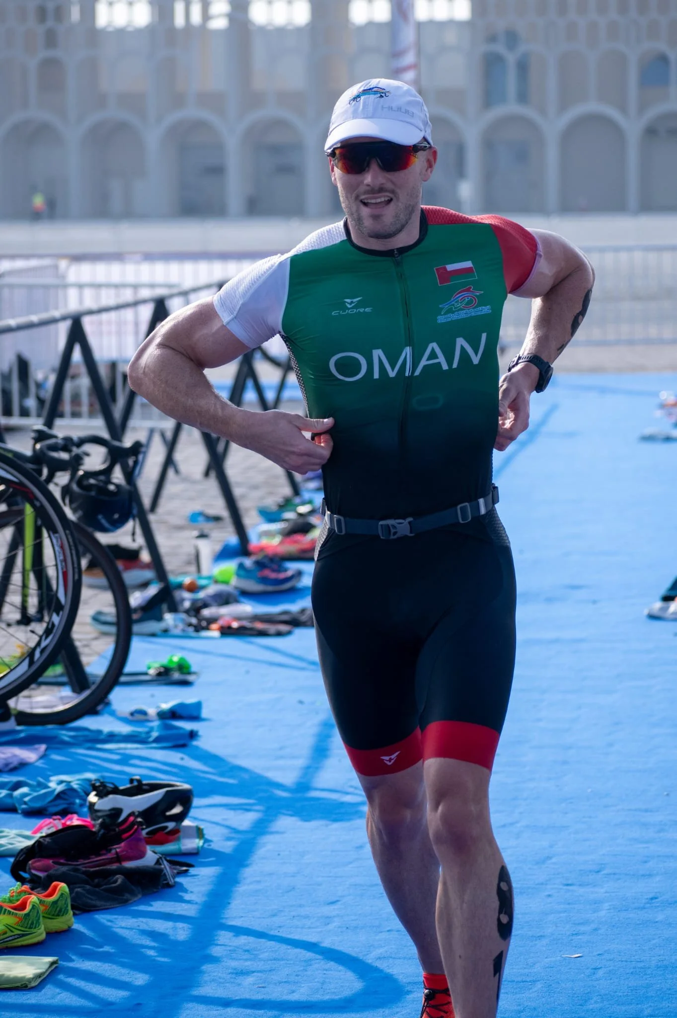 Athlete in green, black, and red triathlon suit running during a race with gear and shoes laid out on blue ground.