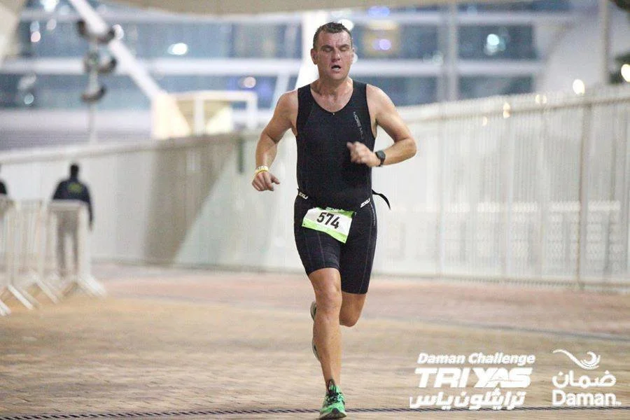 Male runner participating in the Daman Challenge triathlon race, wearing a black sleeveless top and black shorts, with race bib number 574, running on a track indoors or at a sports complex.