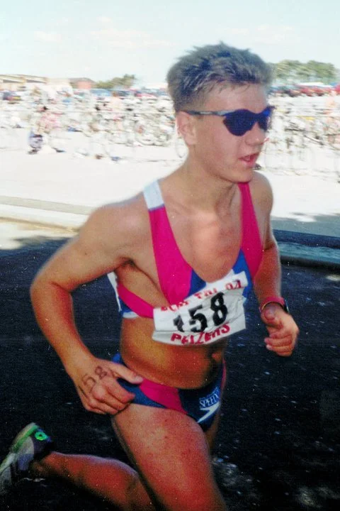 A female athlete running in a race, wearing sunglasses, a pink and blue sports bra and shorts, with a race bib numbered 158, on a track with a crowd in the background.