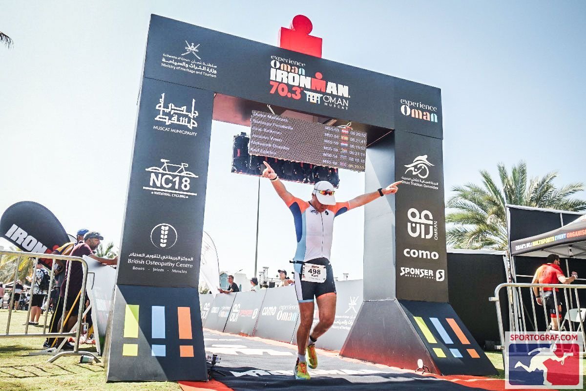 Male athlete crossing the finish line of an Ironman 70.3 race with arms raised in victory, wearing a white cap, sunglasses, and a triathlon suit, with race branding and sponsors visible on the arch and banners.