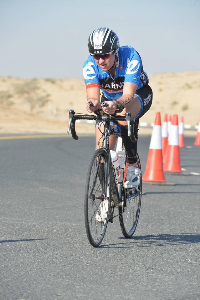 A cyclist in blue race gear and helmet riding a road bike on a paved course marked with orange traffic cones, with sand dunes in the background under a clear sky.
