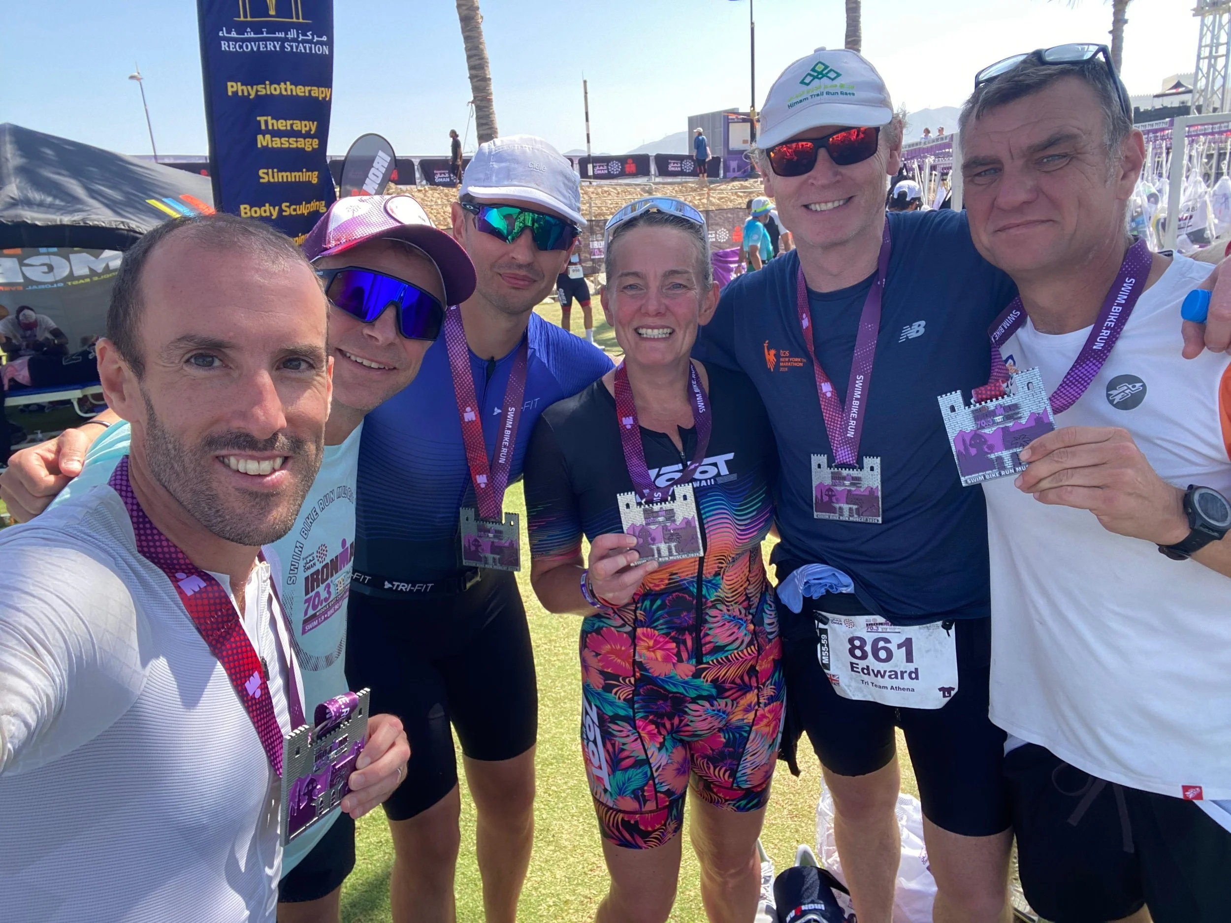 Group of six smiling marathon runners holding medals at a race event, with a blue banner and outdoor tents in the background.