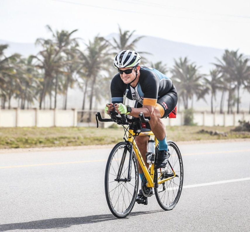 A man riding a yellow road bike on a paved road with palm trees in the background, wearing a helmet, sunglasses, and cycling gear, smiling and leaning forward.