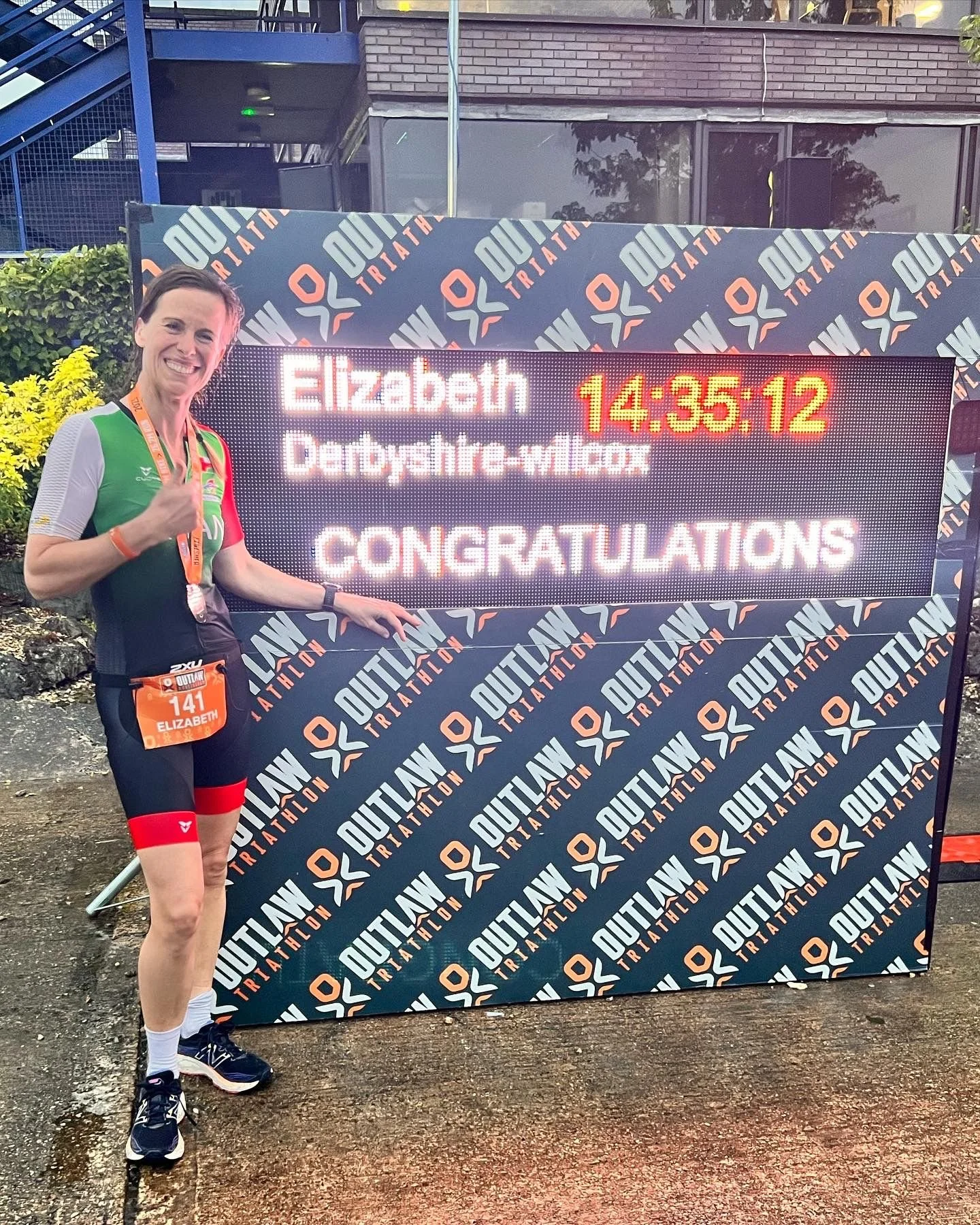 A woman in athletic gear standing next to a sign at a triathlon event, giving a thumbs-up. The sign displays a time of 14:35:12 and congratulates Elizabeth Derbyshe-milcox.