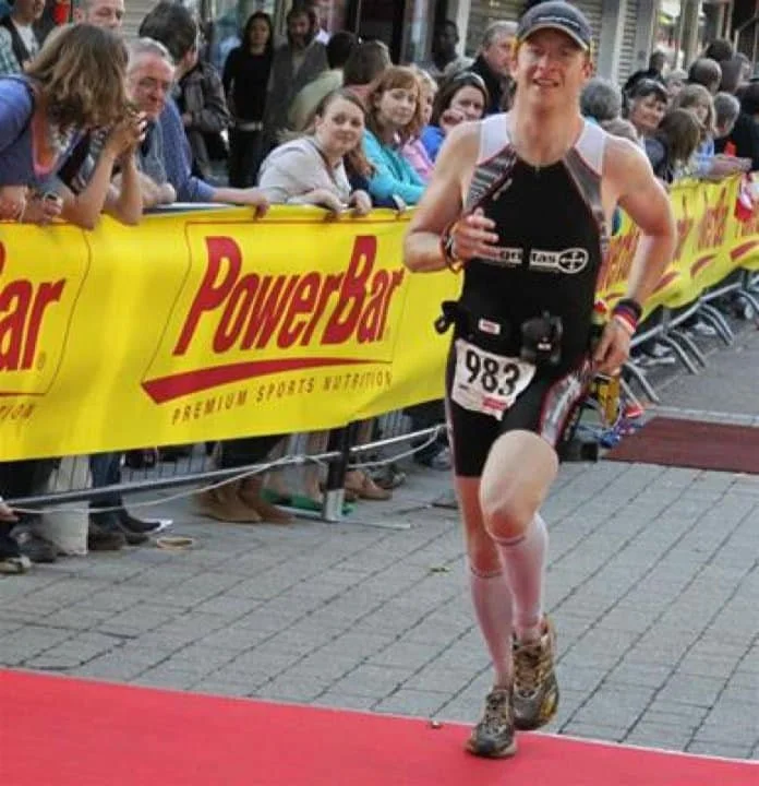 A female athlete crossing the finish line of a race, with spectators cheering behind a yellow banner that reads PowerBar.